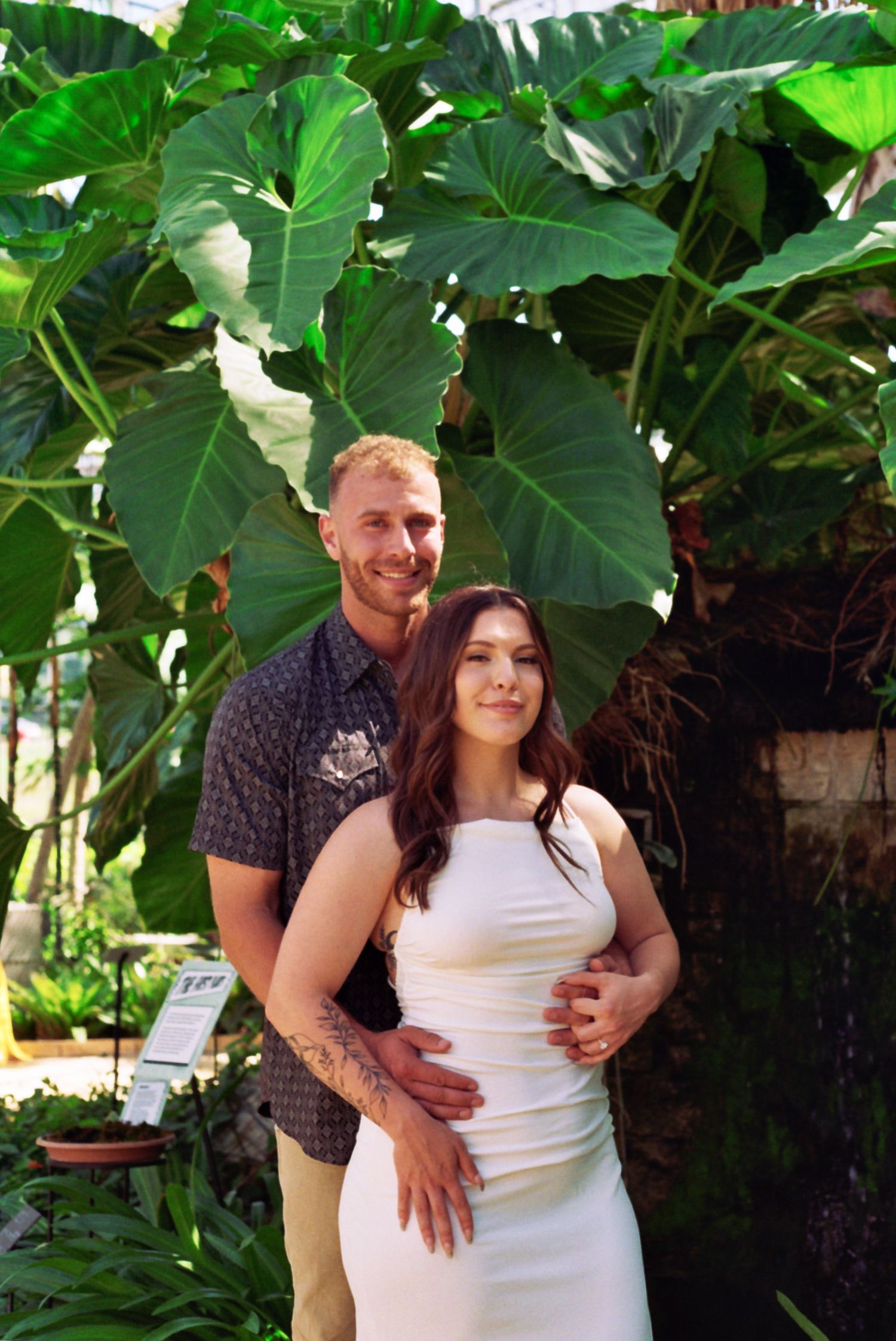 A couple embraces among lush green tropical foliage during a romantic photoshoot in a garden setting.