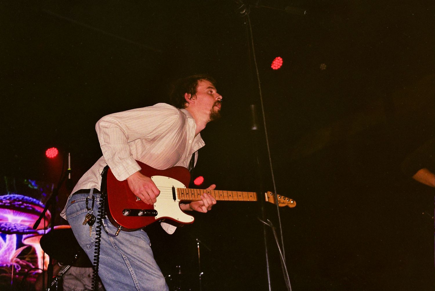 Musician in white shirt bending forward while playing electric guitar during dark concert performance.