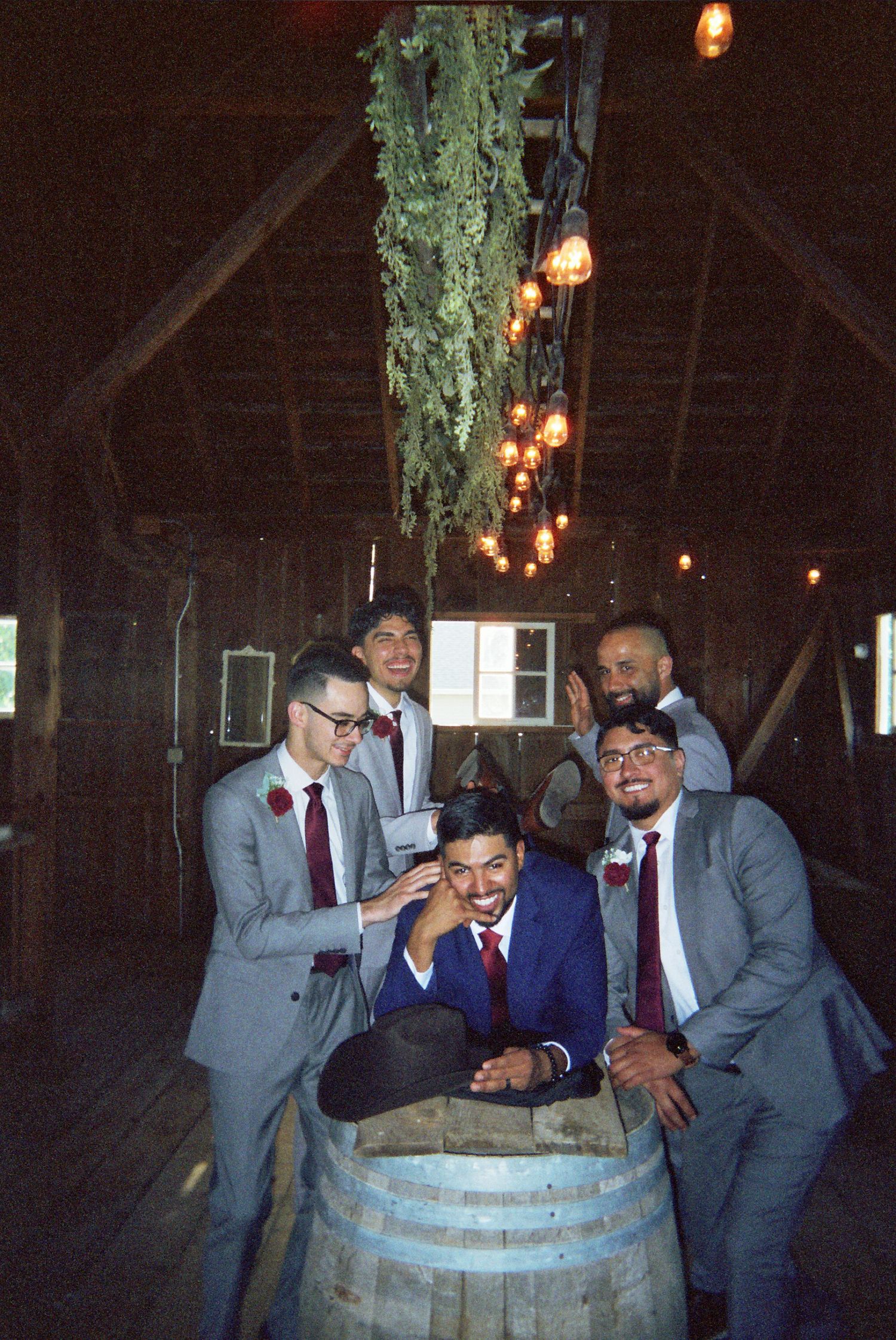 A group in gray suits gather around a wooden barrel inside a rustic barn with hanging lights and exposed beams.