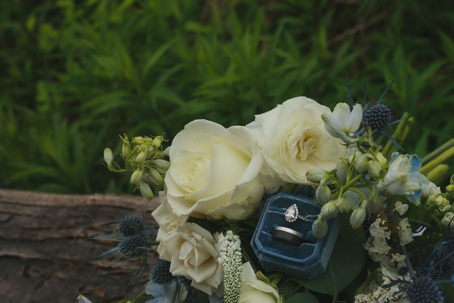 Wedding ring in navy blue velvet box with white roses and greenery on rustic wooden surface in garden setting.