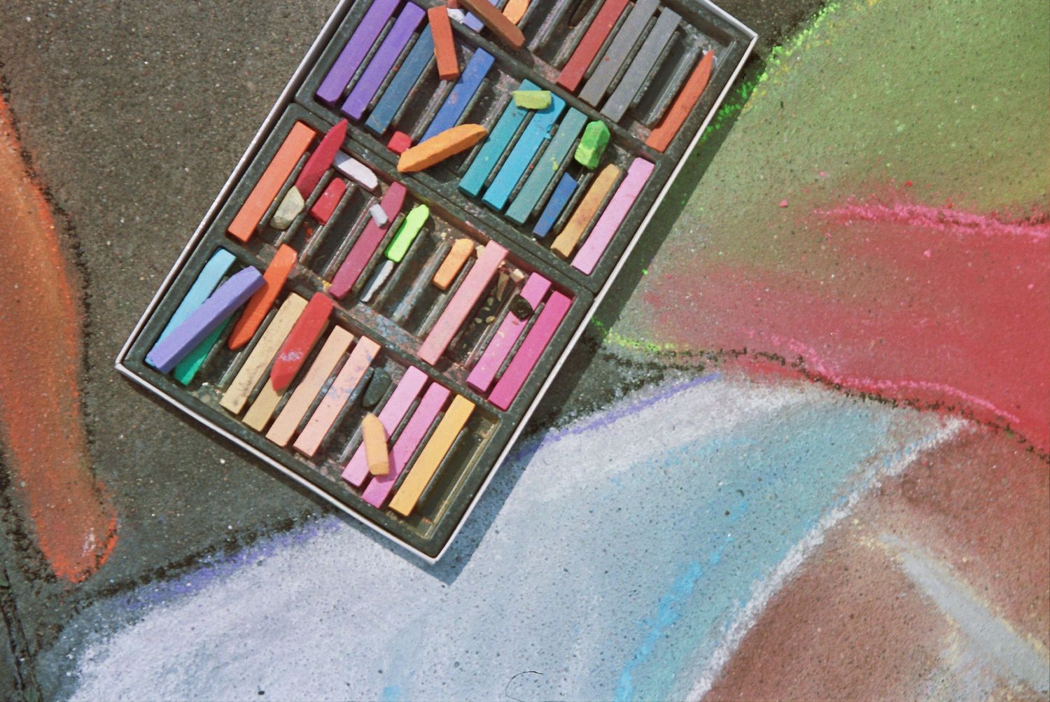 Colorful chalk pastels in a black tray beside vibrant sidewalk chalk art on pavement.