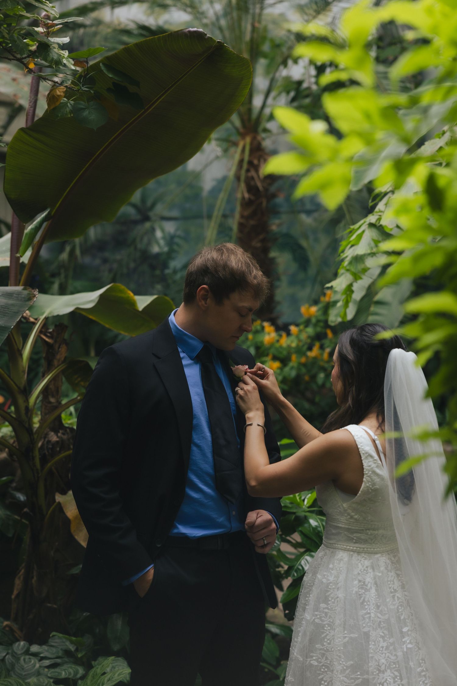 Bride adjusting groom's boutonniere in lush tropical greenhouse wedding setting with green foliage backdrop.