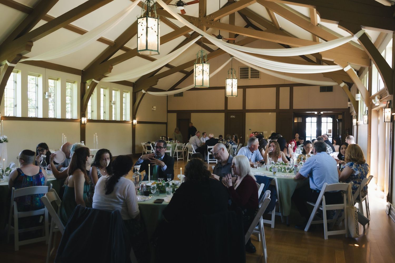 Elegant wedding reception in a rustic hall with wooden beams, draped fabric, and guests dining at round tables.
