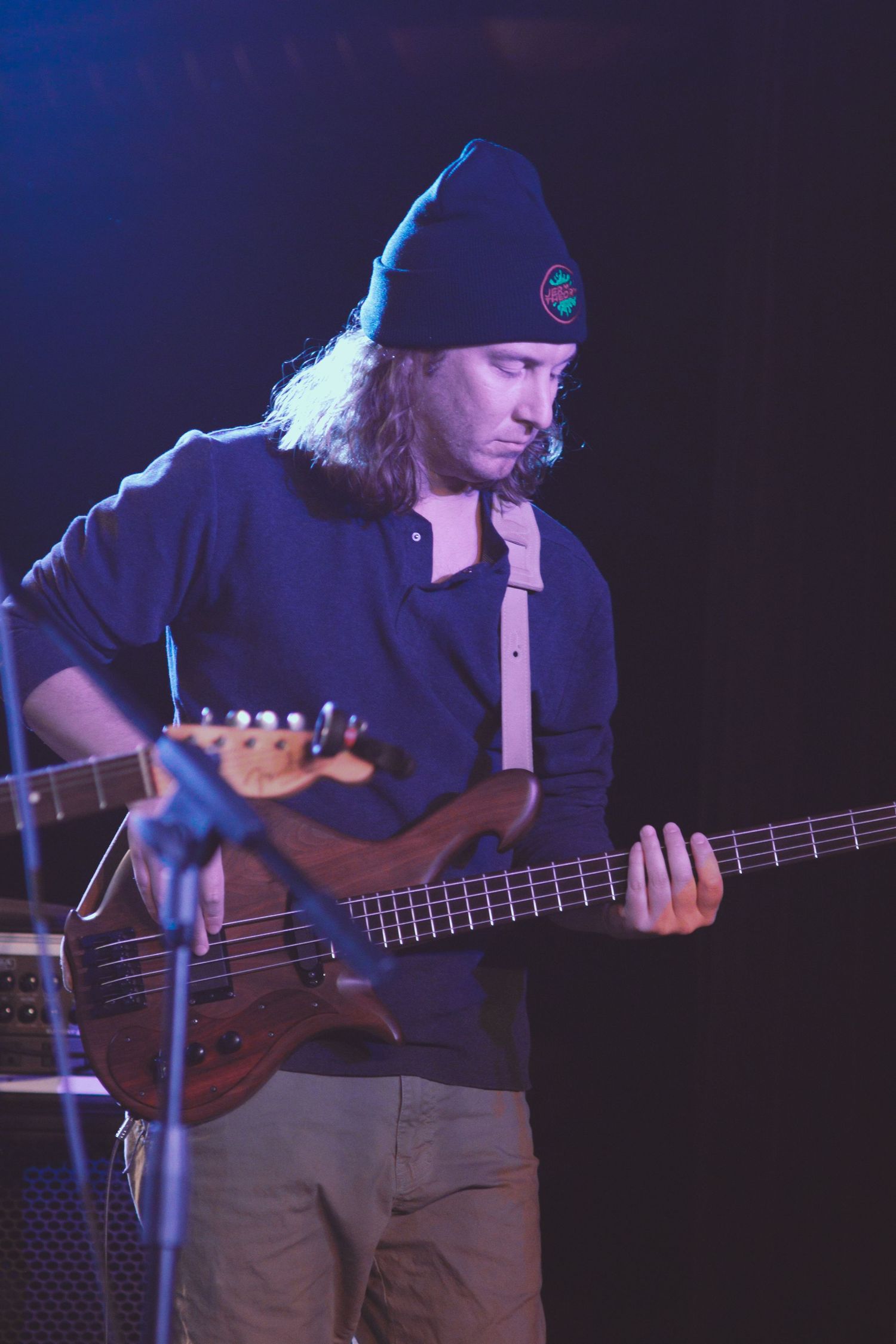 Bass guitarist performs on stage wearing dark clothing and a beanie under blue stage lighting.