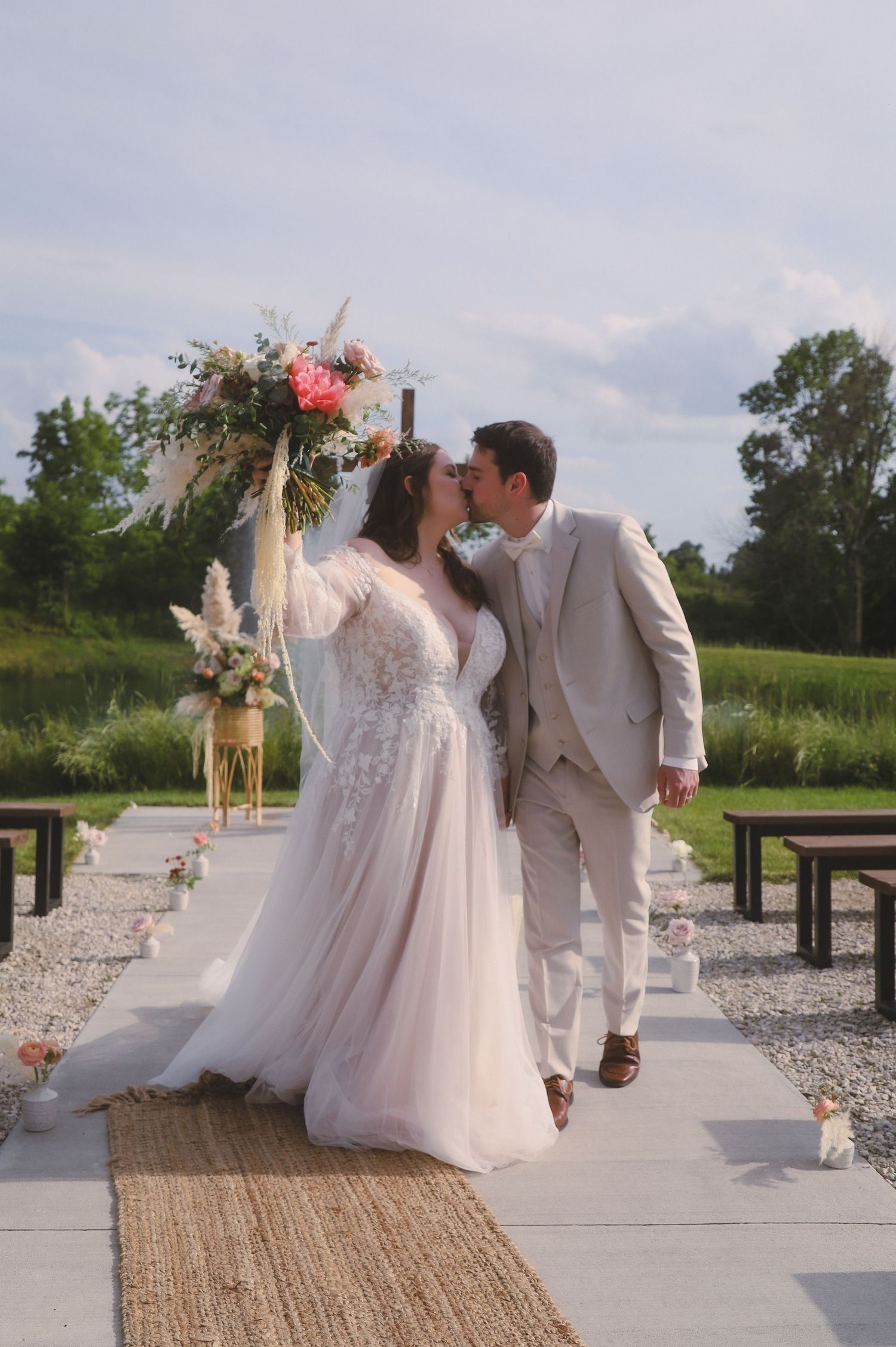 Romantic outdoor wedding ceremony with couple embracing under floral arch on stone path with rustic benches and decor.