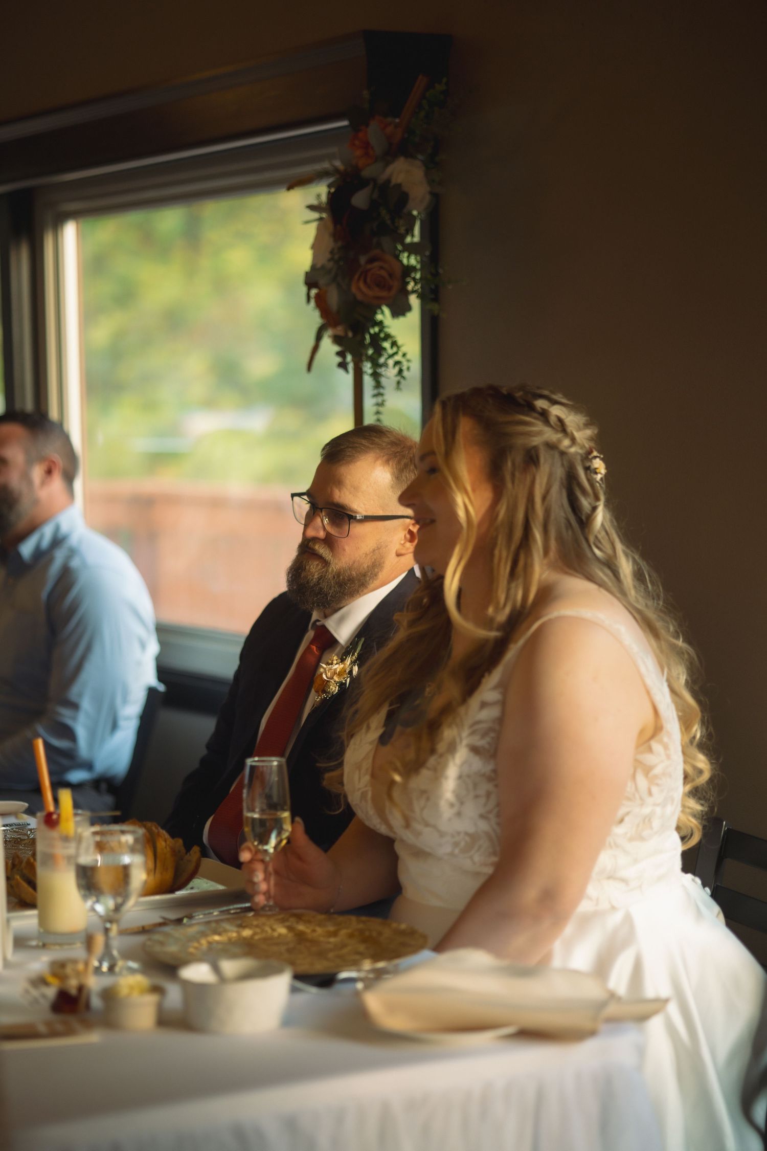 Wedding couple seated at reception table with floral decorations and window light.