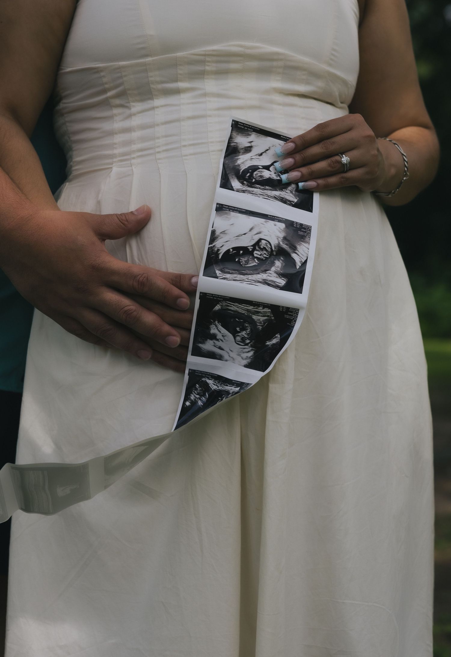 Pregnant woman in white dress holding ultrasound photos against baby bump with partner's hand resting on belly.