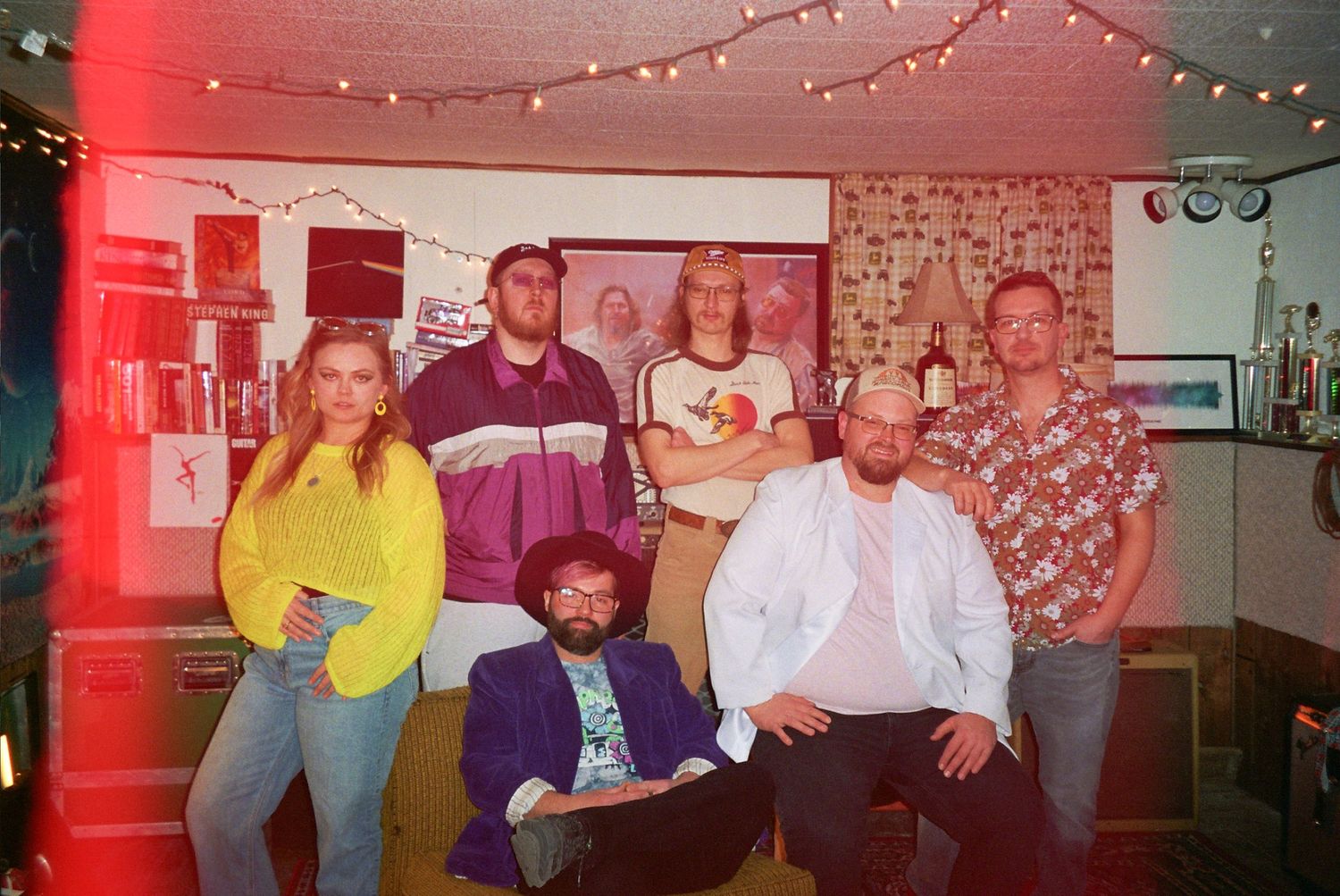 A group of seven friends posing together in a retro-styled room with string lights and colorful outfits.