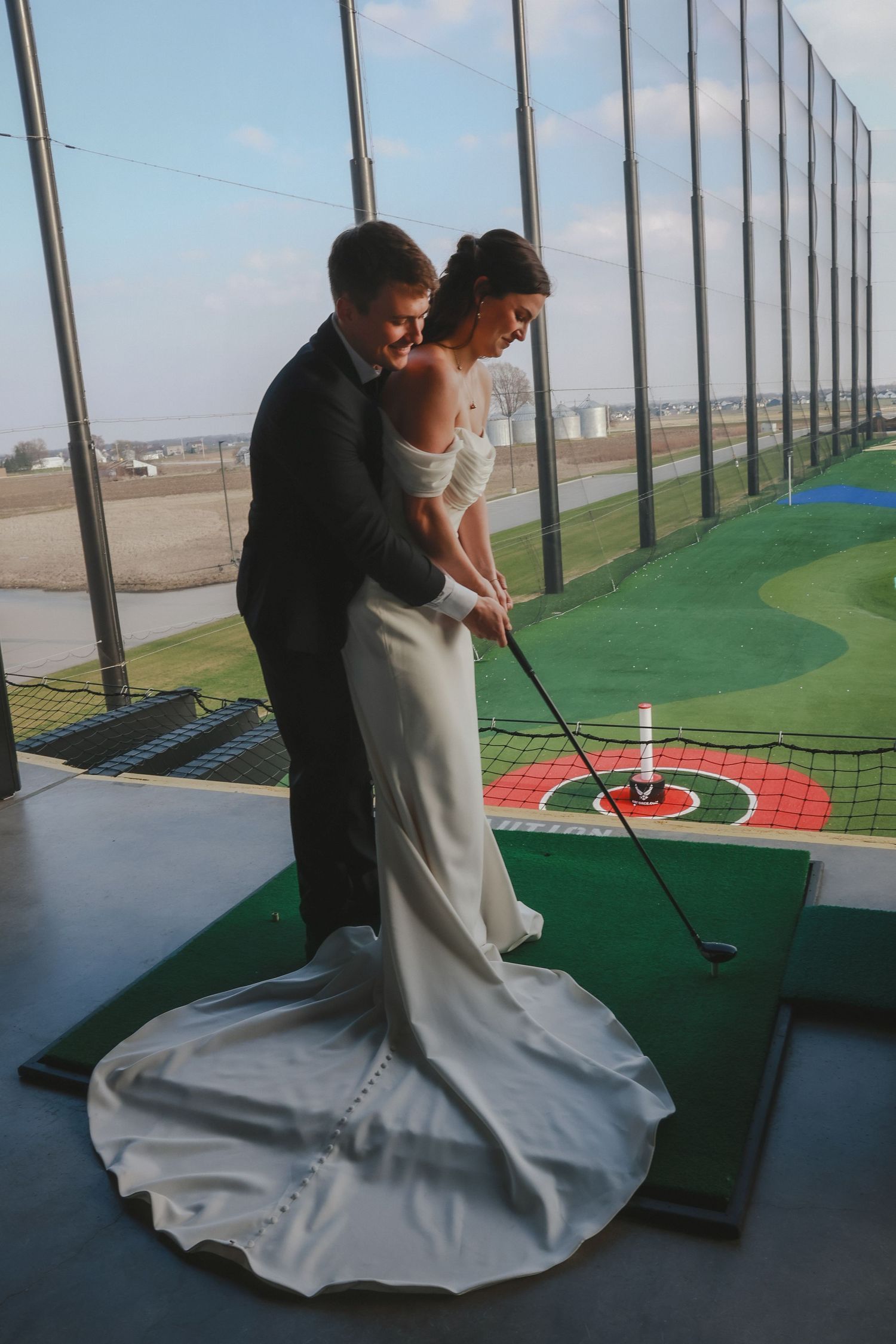 A couple in formal wedding attire practicing their golf swing together at a driving range.