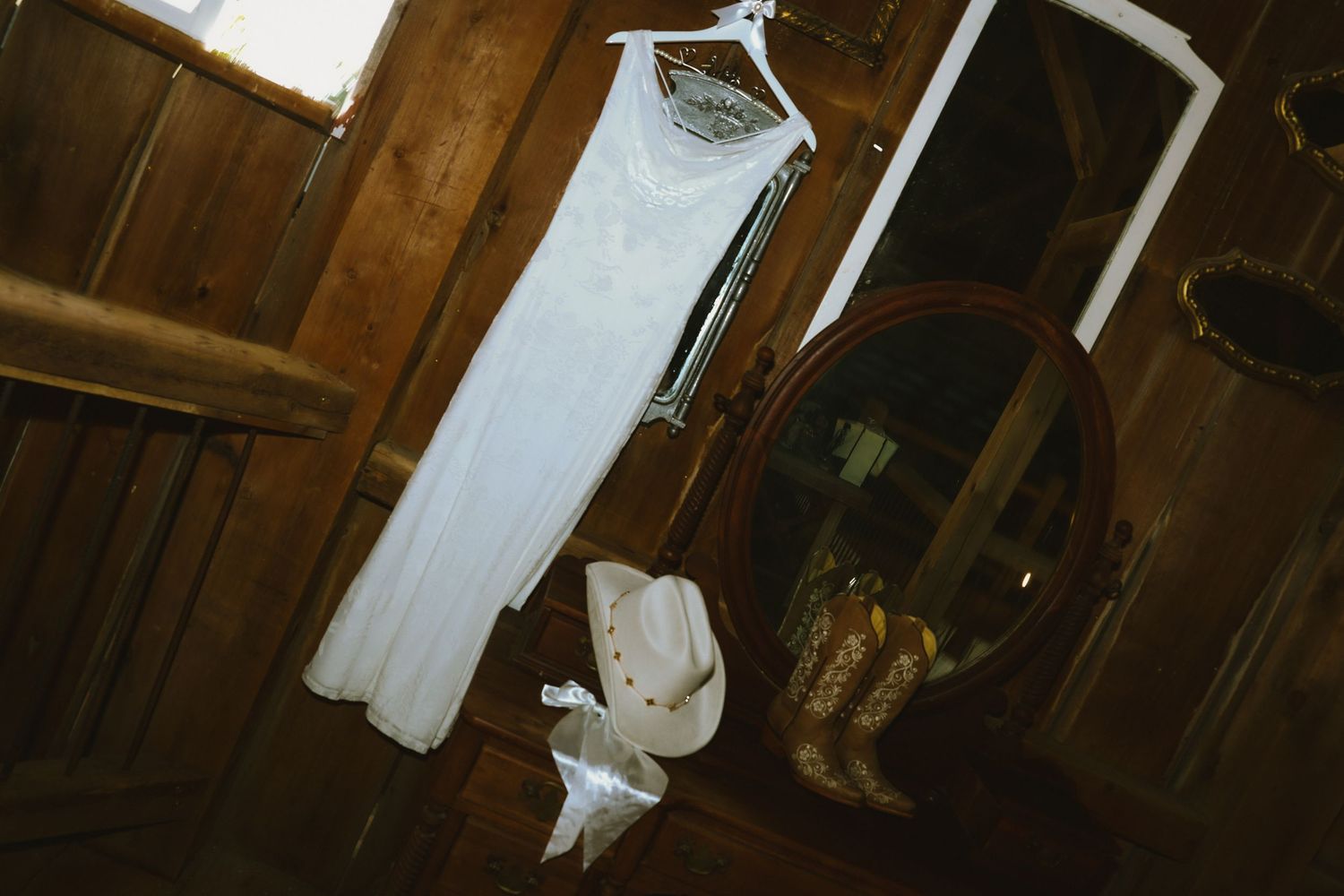 A white wedding dress and pair of cowboy boots hang in a rustic wooden room, creating a country wedding aesthetic.