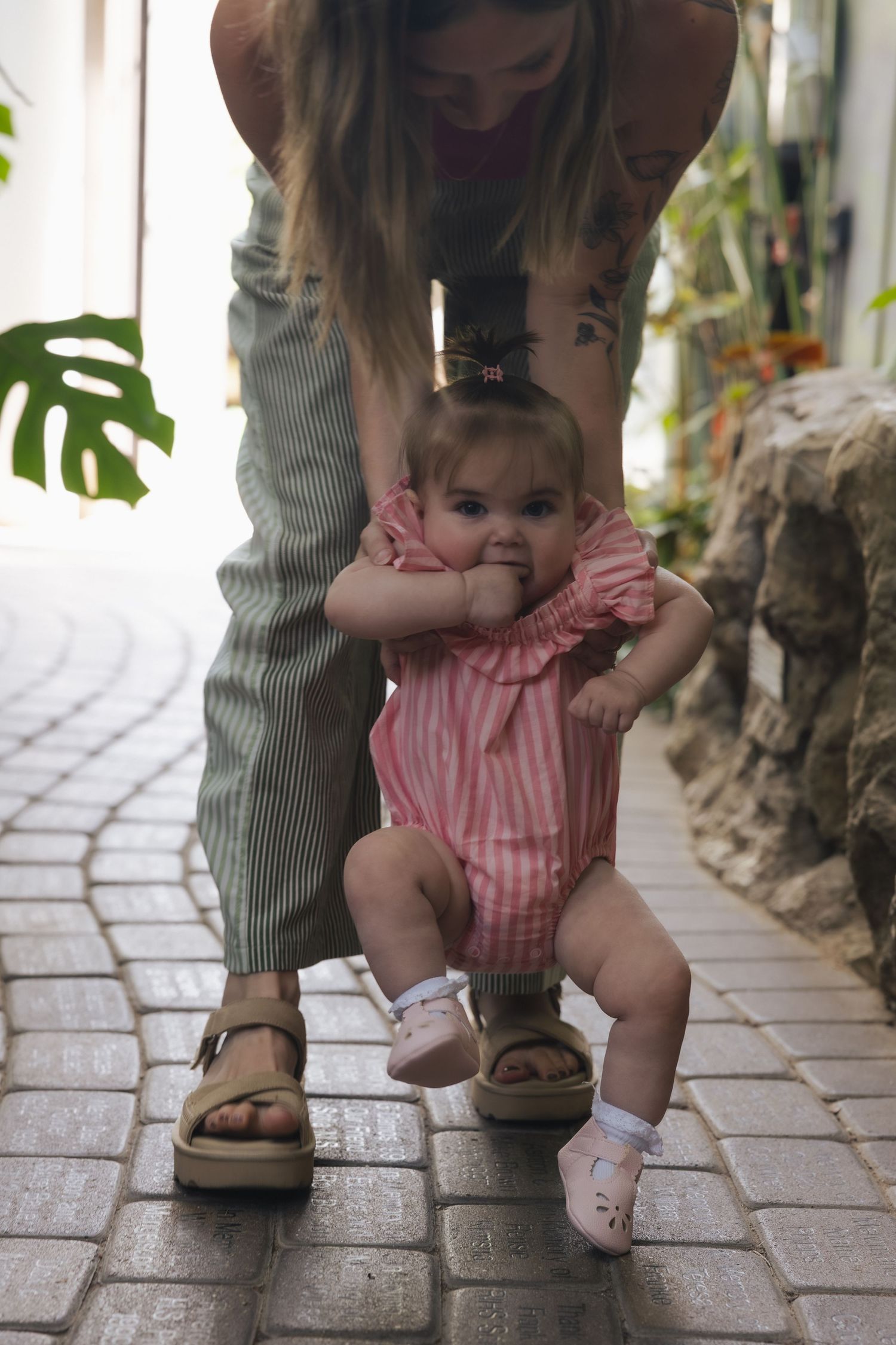 Mother helping baby girl in pink romper take first steps on cobblestone path outdoors.