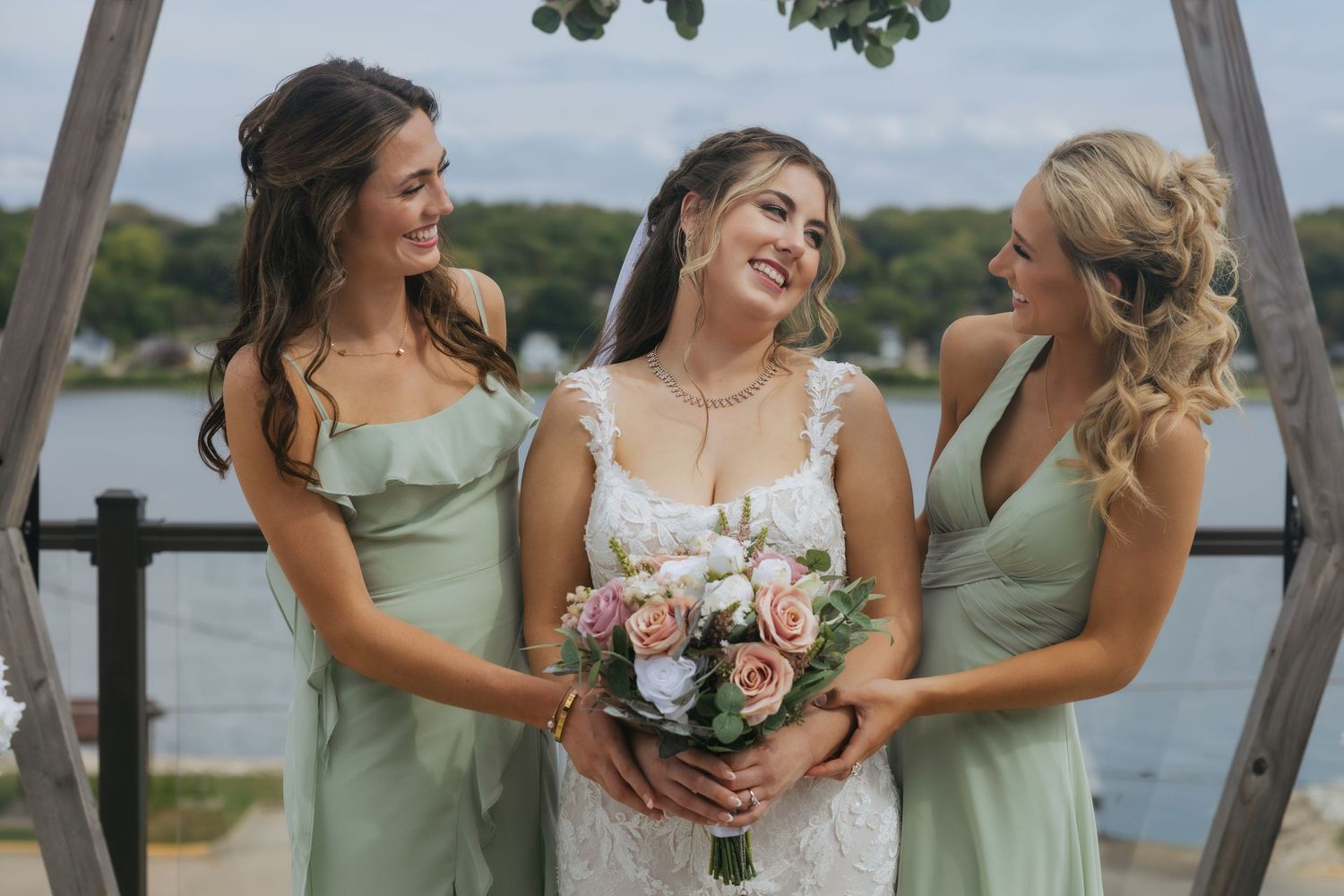 Bridesmaids in sage green dresses share joyful moments during outdoor wedding photo session by the water.