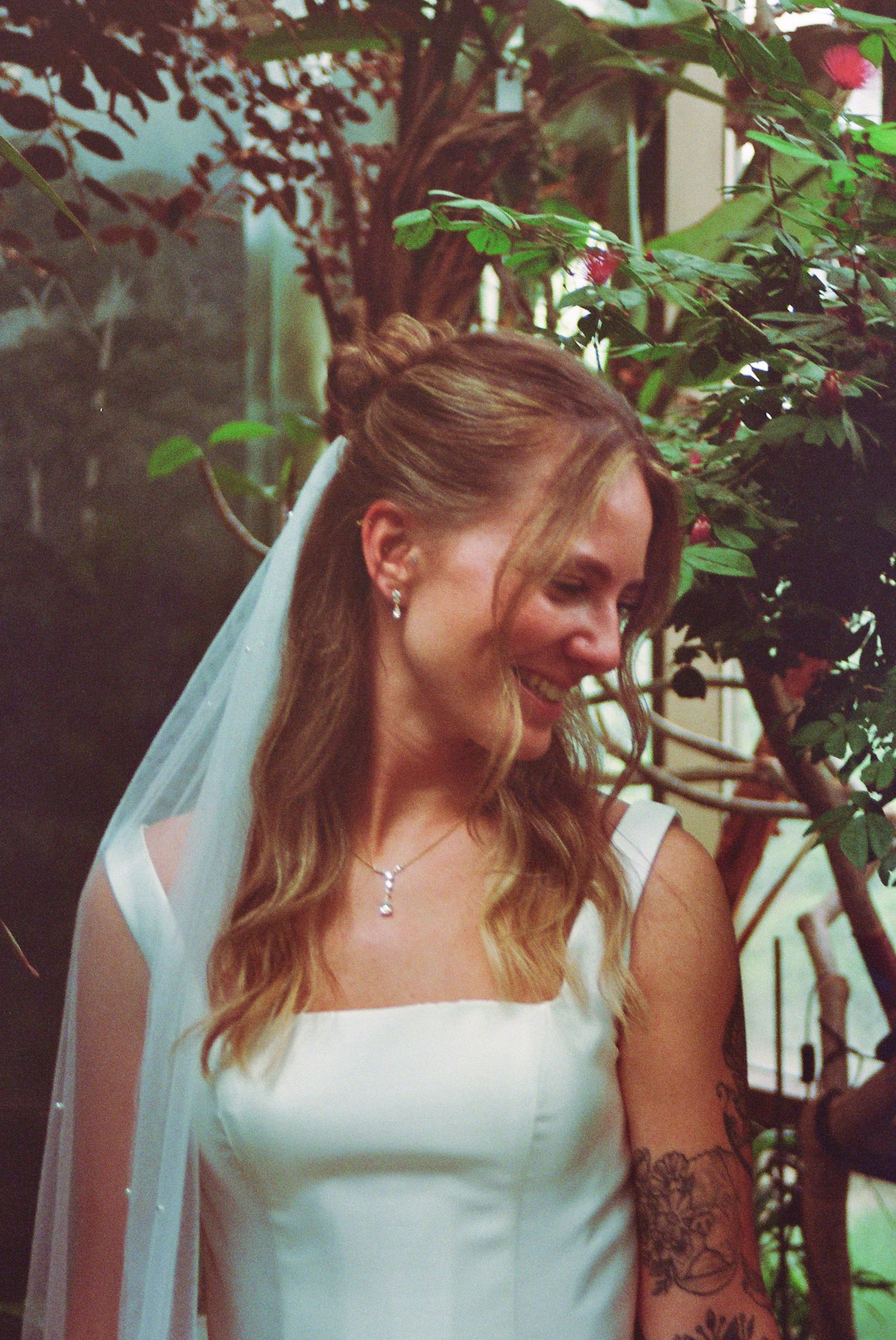 Smiling bride in white gown and veil with tattoo, standing among lush green foliage outdoors.