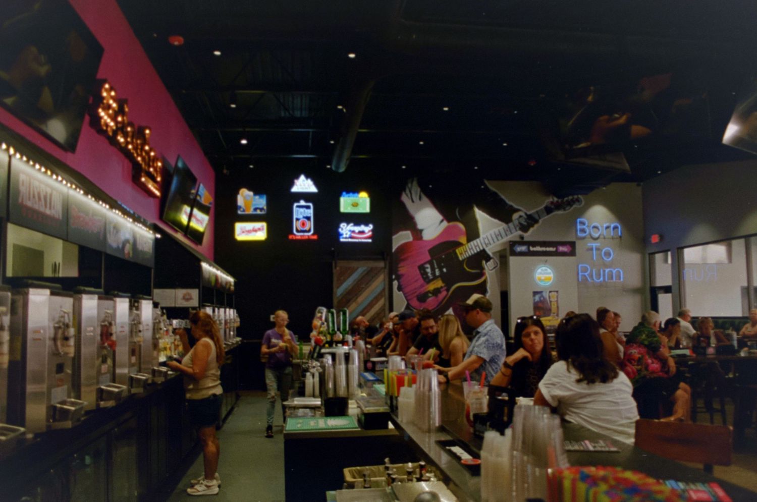 Busy bar interior with neon signs, bartenders serving drinks, and patrons gathered along a colorful counter.