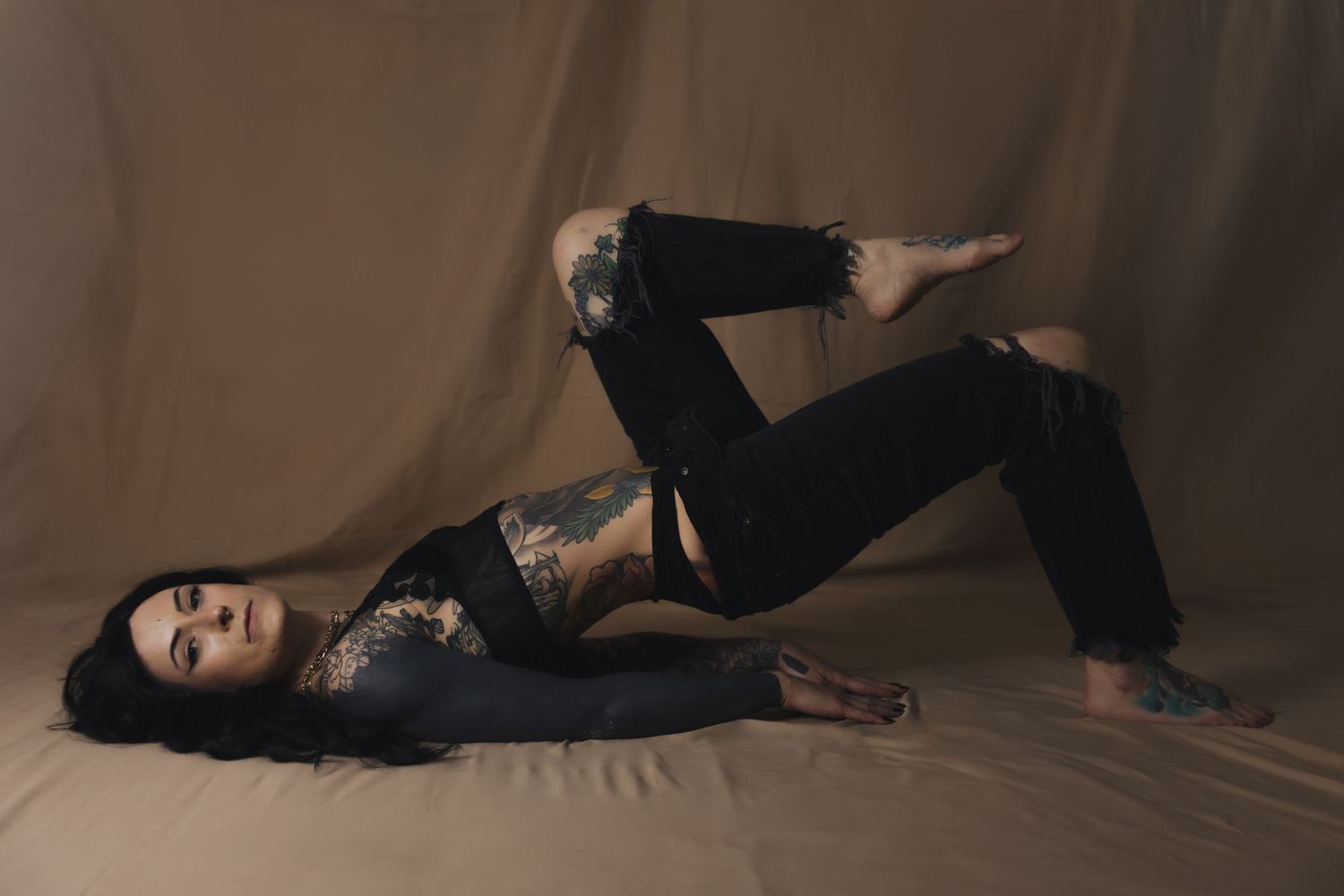 Tattooed woman in black outfit posing gracefully on a tan studio backdrop, showcasing body art and fashion.