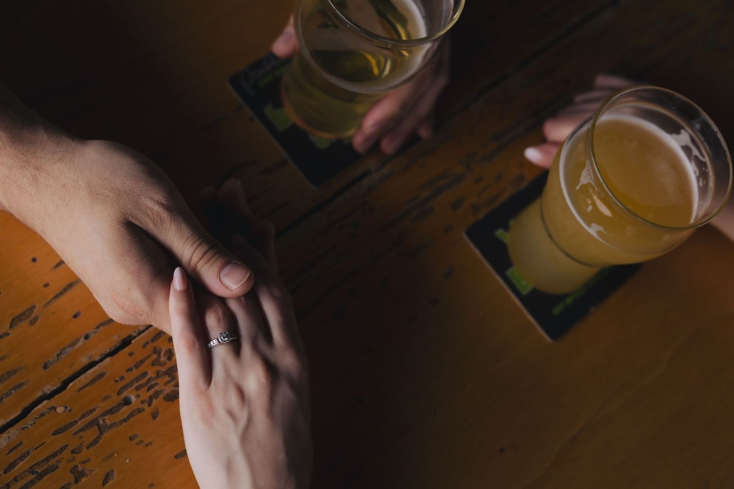 Two people hold hands on a wooden table while sharing glasses of cold beer.