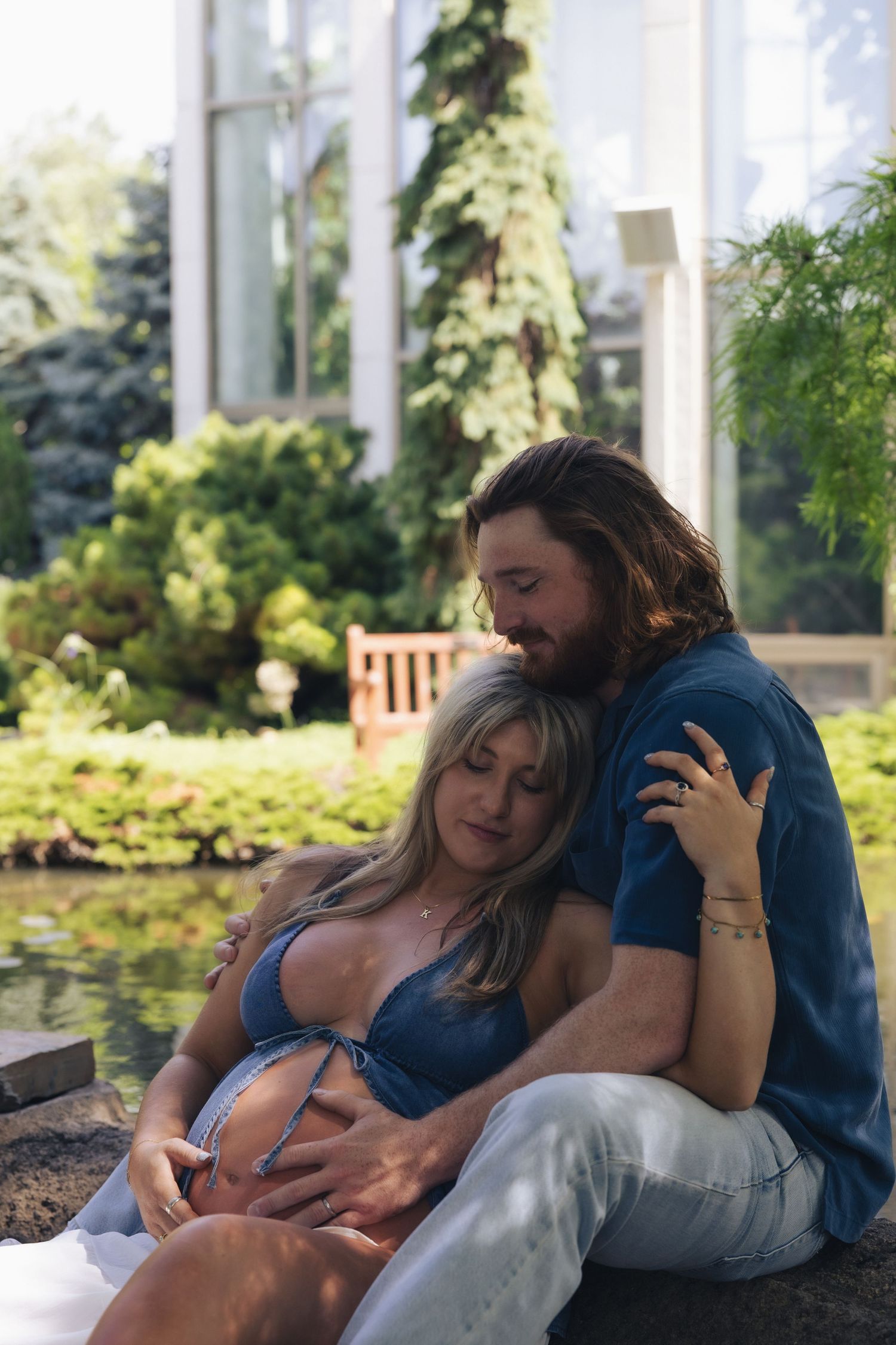 Couple sitting outdoors by a pond, pregnant woman in blue bikini top cradled by partner in lush garden setting.