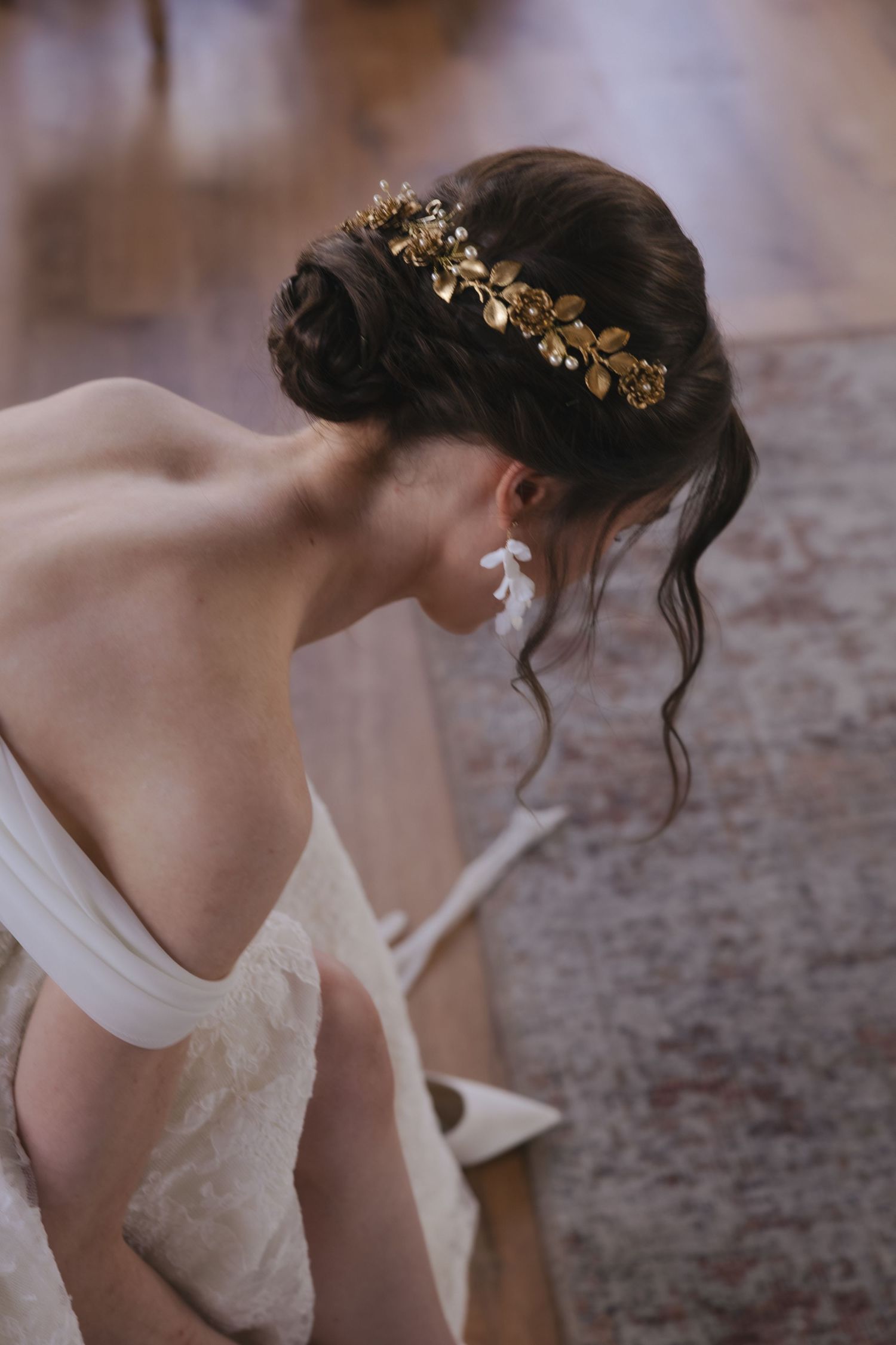 Bride in lace gown with gold floral crown and white leaf earring, bending down, elegant updo hairstyle.