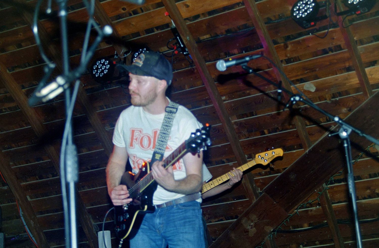 Male guitarist performing on stage in a rustic barn venue, wearing a cap and Folk Nation t-shirt.