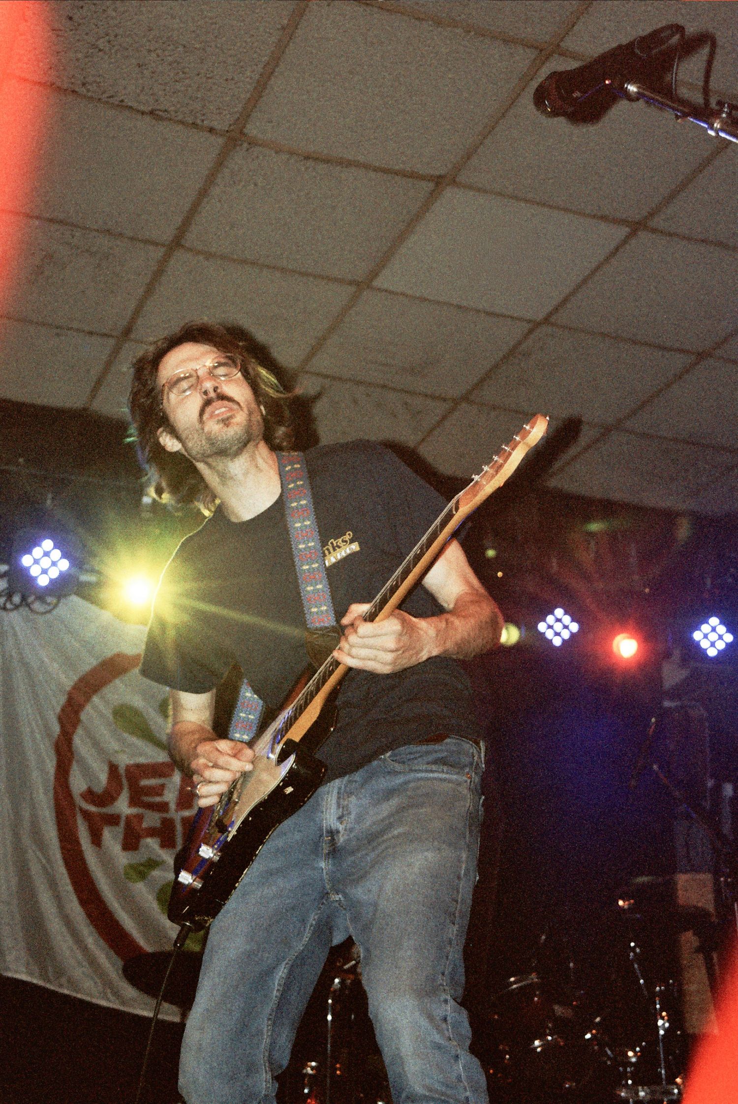Male guitarist performing on stage with colorful concert lights, playing electric guitar in casual jeans and black t-shirt.