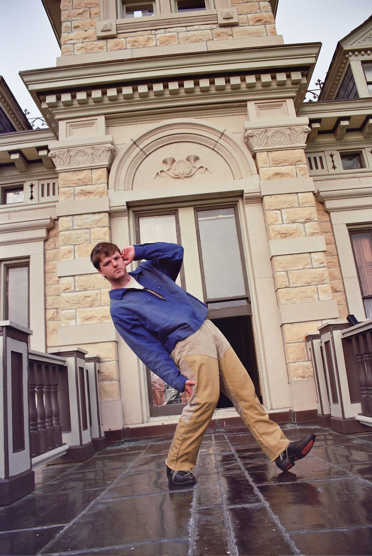 Young man in blue hoodie and khaki pants striking a dynamic pose on wet pavement outside a historic stone building.