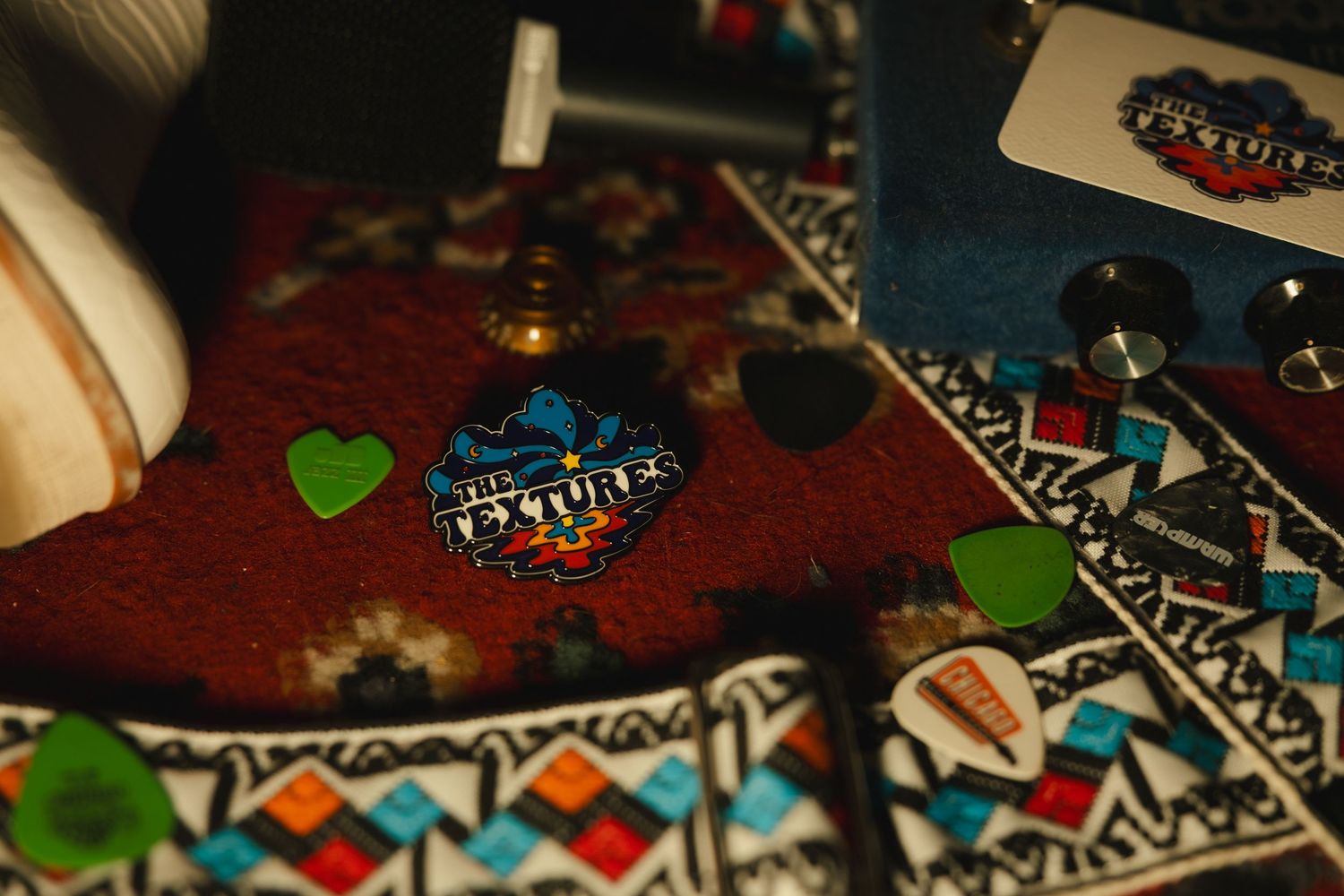 Close-up of colorful guitar picks, a green heart, and a band pin on a decorative patterned surface.