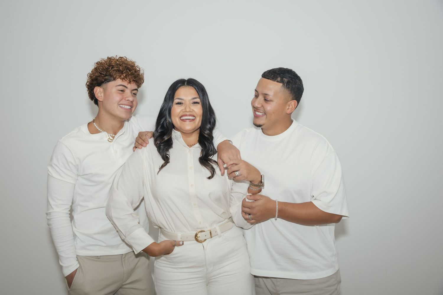 Three people in white outfits smiling and posing together against a light gray background.