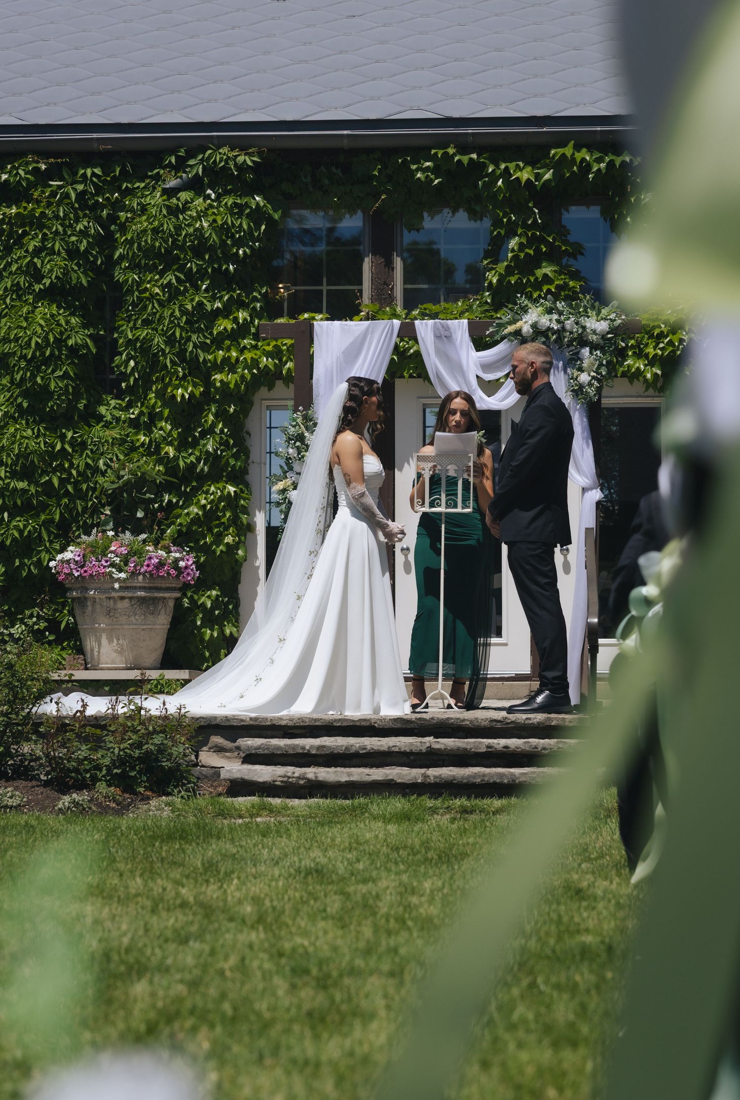 Outdoor wedding ceremony with bride in white gown and groom at floral arch beside officiant on sunny day.