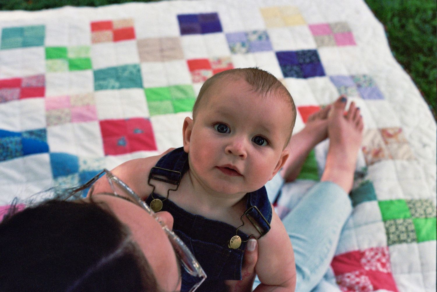 A young child in denim overalls laying on a colorful patchwork quilt outdoors in the grass.