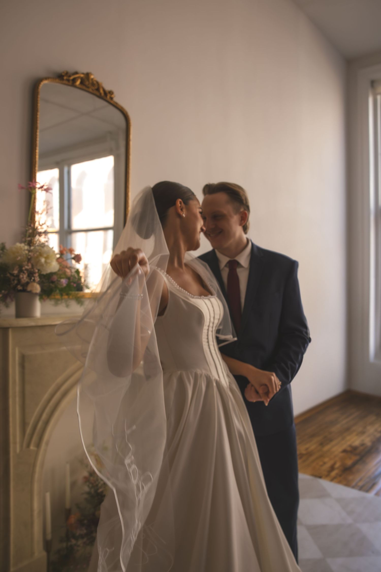 A couple in an elegant room with an ornate gold mirror share an intimate moment on their wedding day.