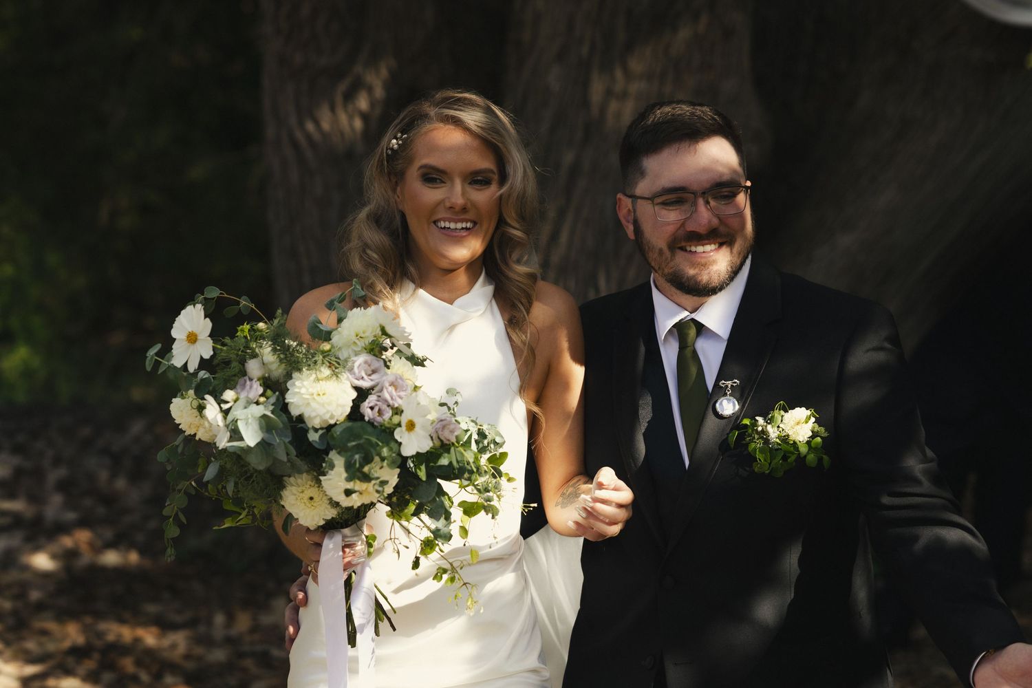 A couple poses together in formal wedding attire holding bouquets of white flowers in an outdoor garden setting.
