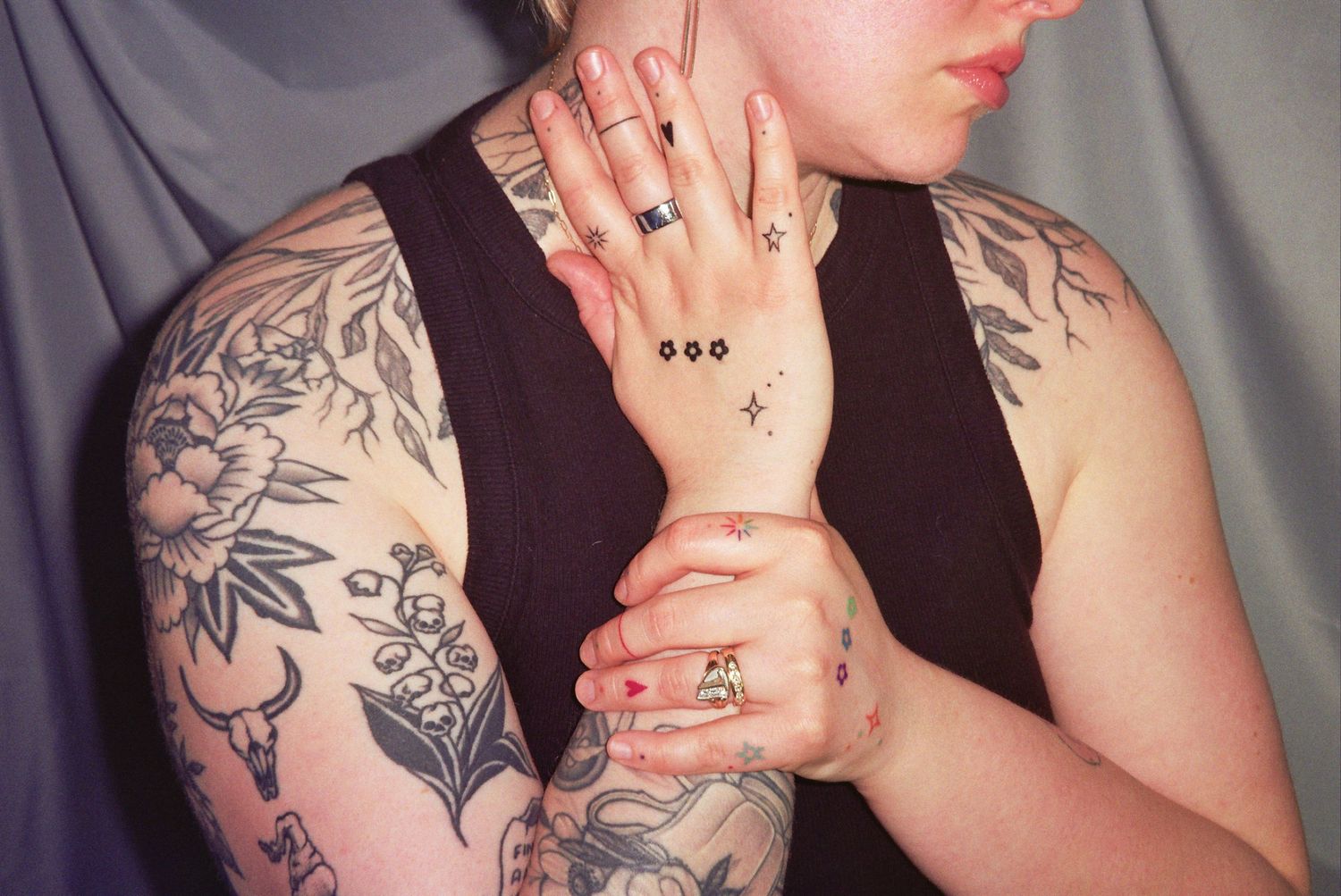 Close-up of tattooed arms with floral sleeve tattoo, rings, and small hand tattoos on a person in a black tank top.