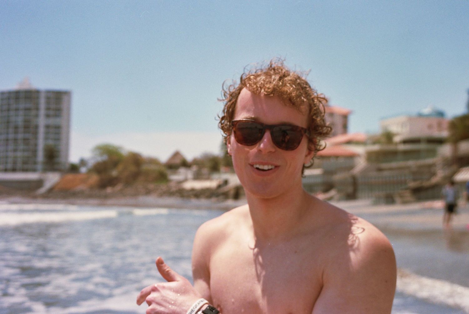 Smiling shirtless man with curly hair and sunglasses at a sunny beach with buildings in the background.
