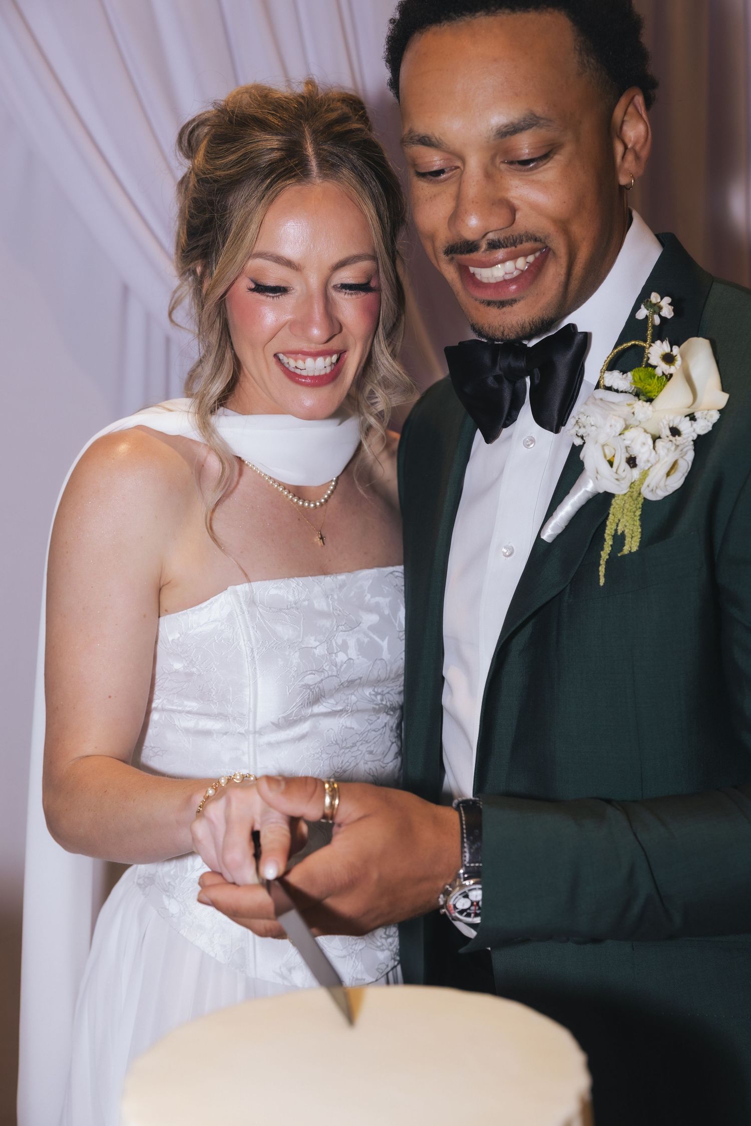 Bride and groom smiling while cutting their wedding cake, bride in white gown, groom in green tuxedo with boutonniere.