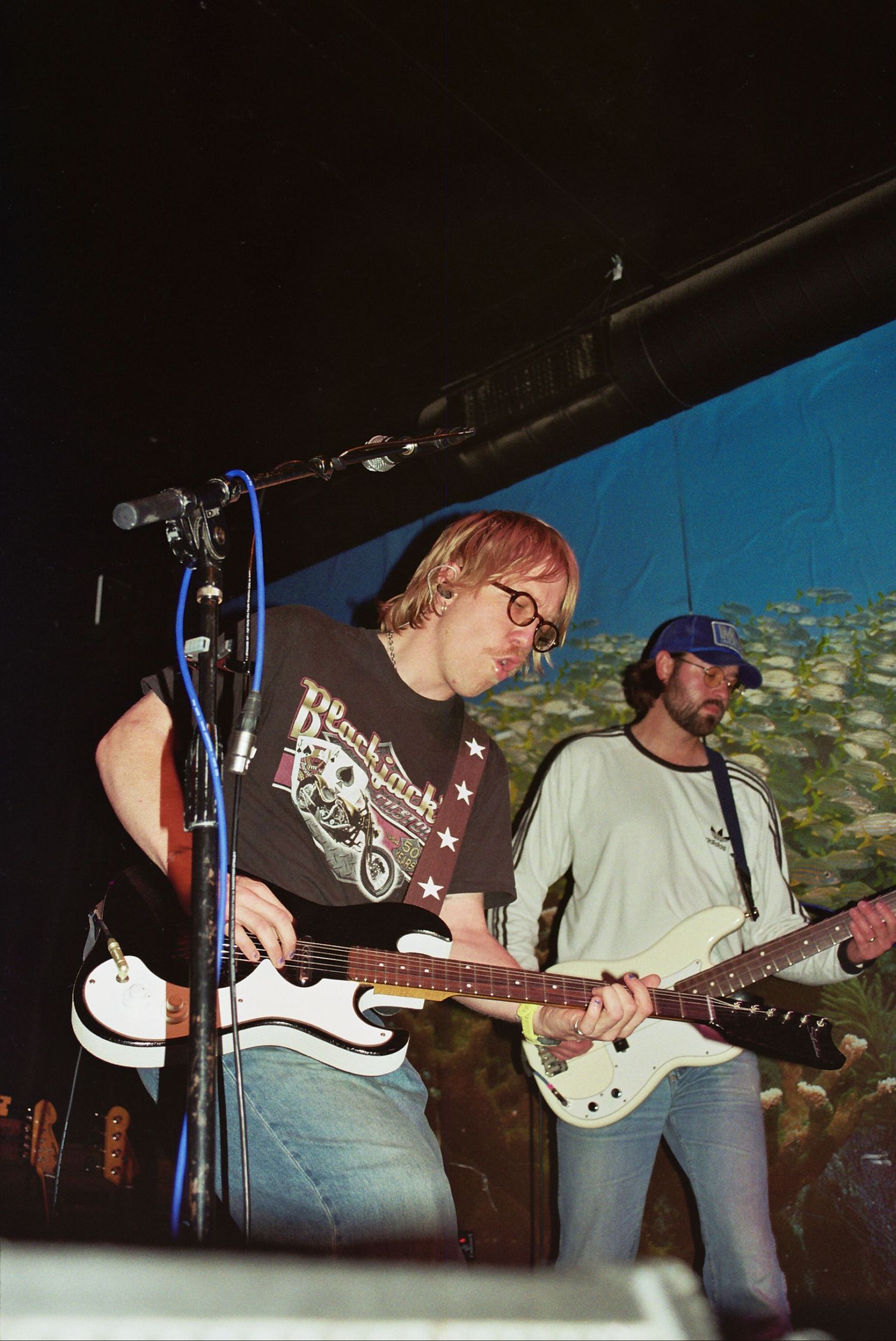 Two guitarists perform on stage, one with pink hair wearing a black tee, the other in a gray shirt and blue cap.