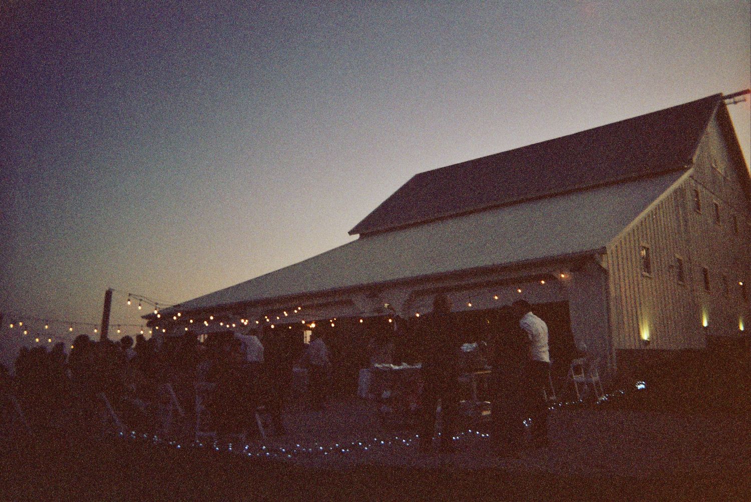 A rustic barn illuminated with string lights at dusk creating a romantic outdoor event setting.