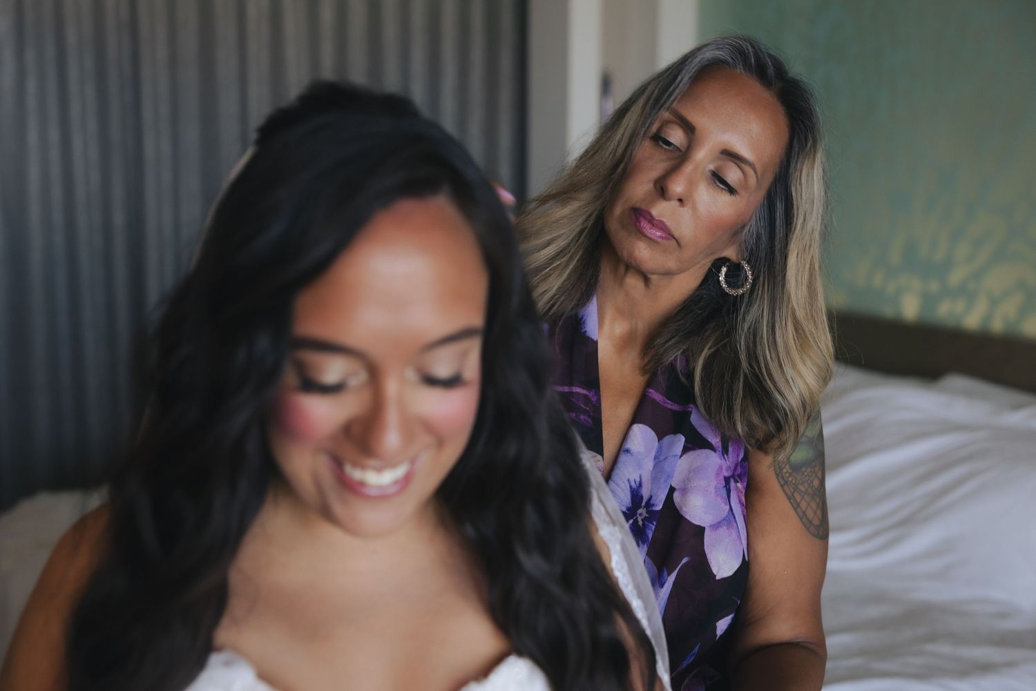 A bride smiles as an older woman helps her prepare for her wedding day while seated on a white bed.