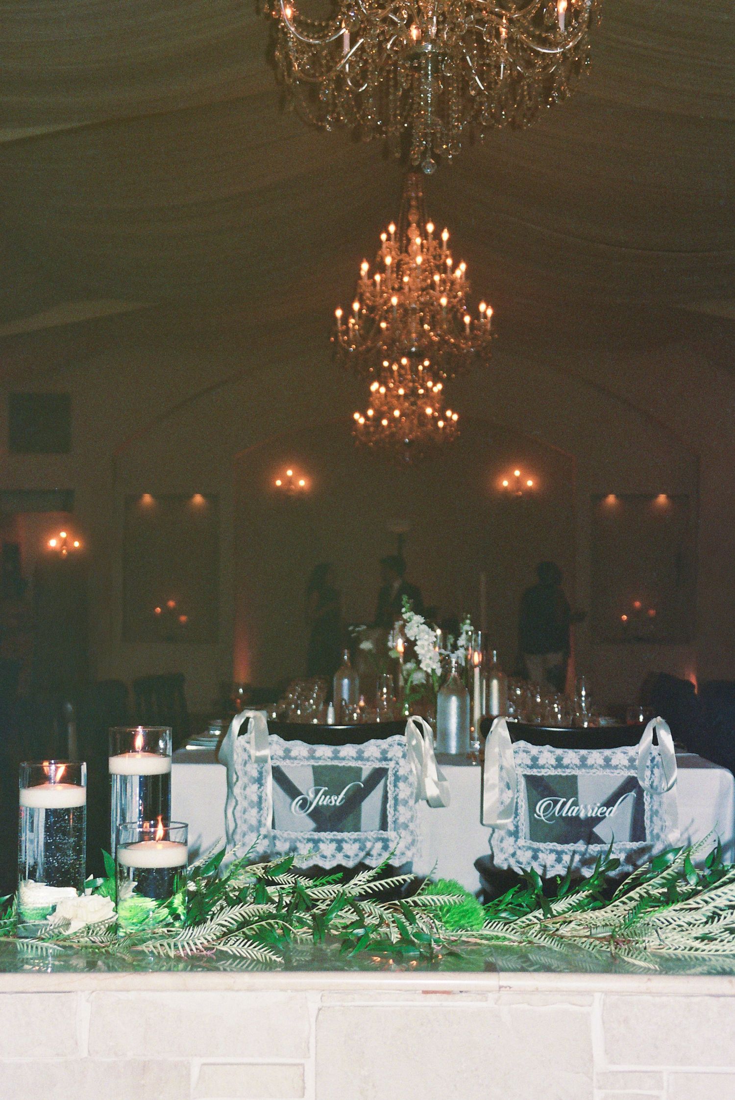 Elegant wedding reception sweetheart table with Just Married signs, chandelier, candles, and greenery decor.