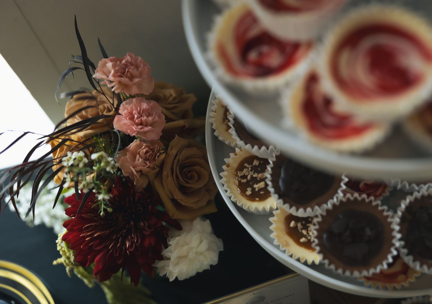 Floral arrangement with roses and carnations beside a plate of fruit tarts and chocolate cupcakes on a dark table.