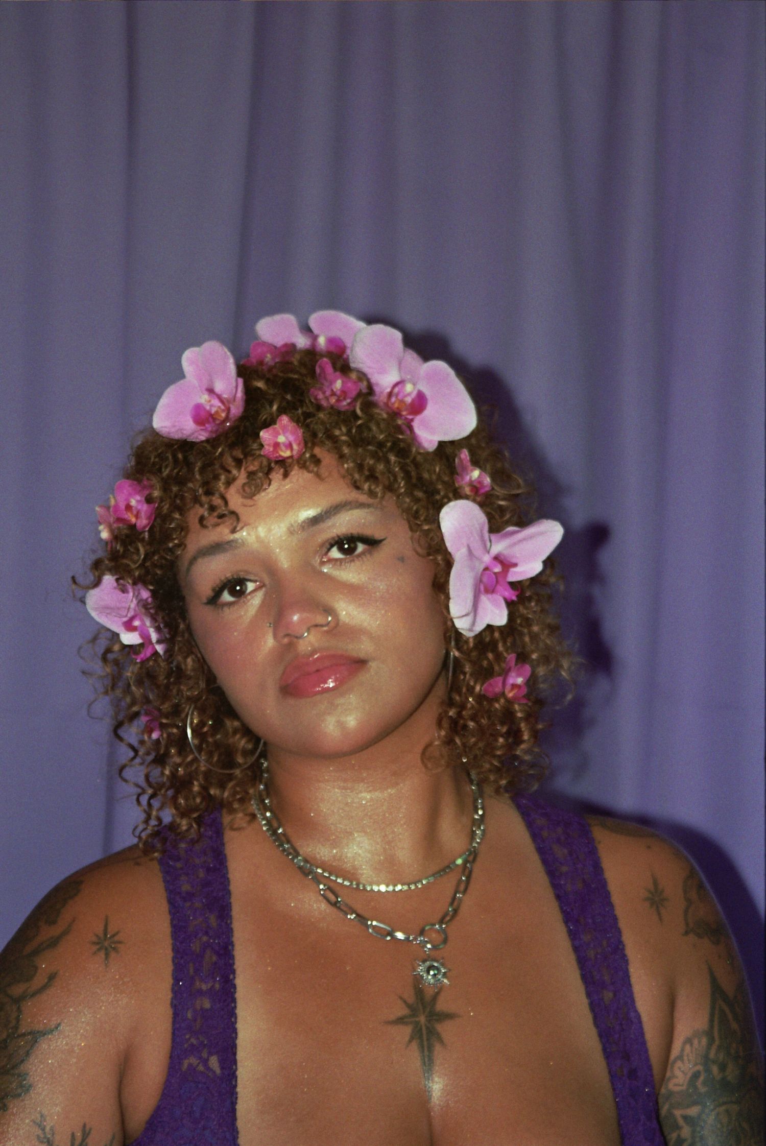 Woman with curly hair adorned with pink orchid flowers wearing purple lace top and layered necklaces with tattoos.
