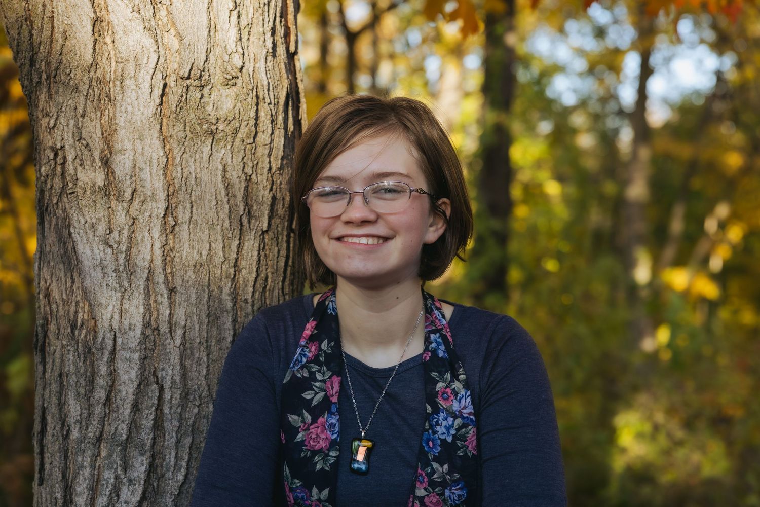 Smiling young woman with glasses leaning against a tree in autumn forest with colorful foliage.