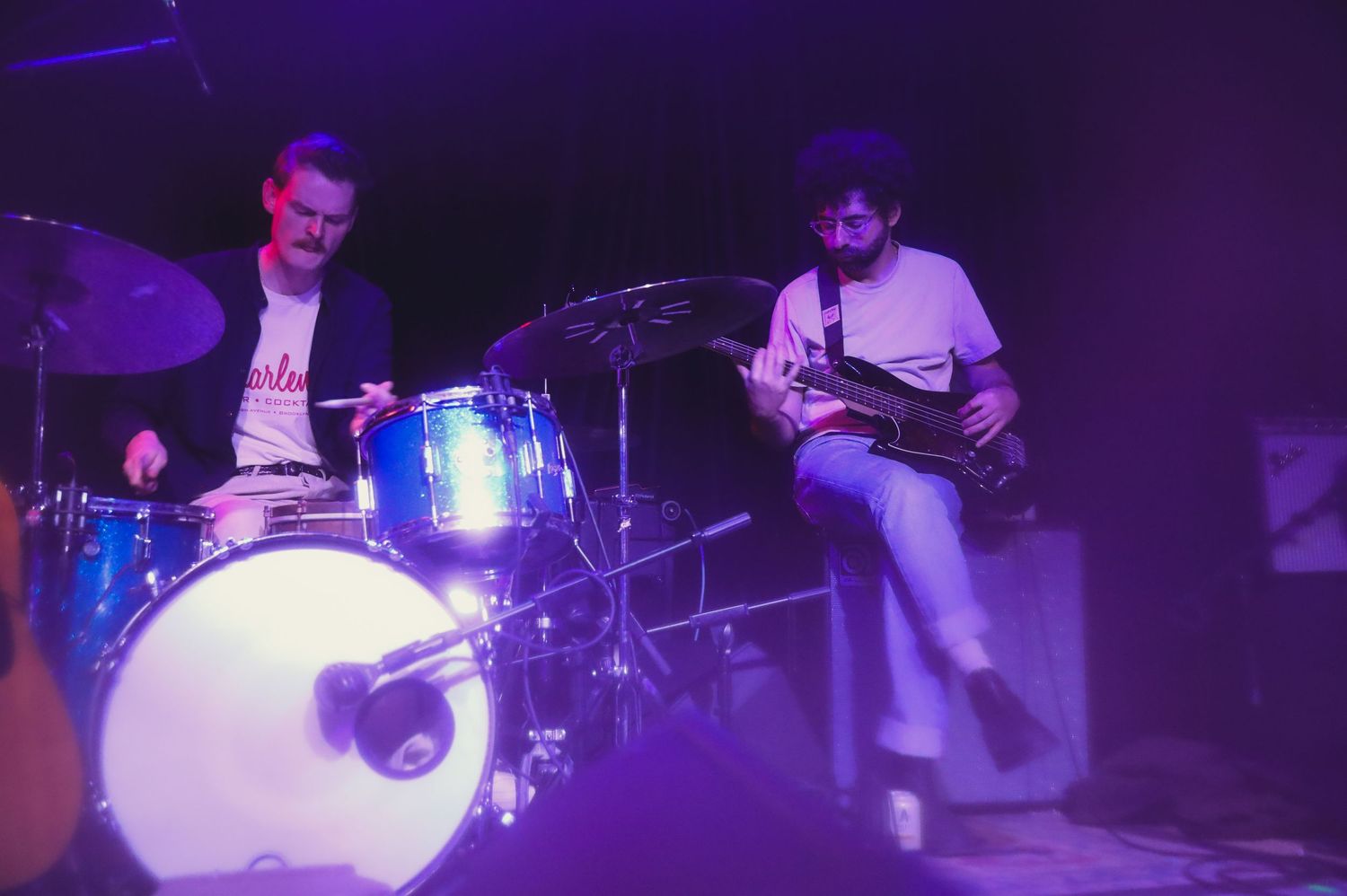 Band performing on dimly lit stage with drums and instruments under blue and purple stage lighting.