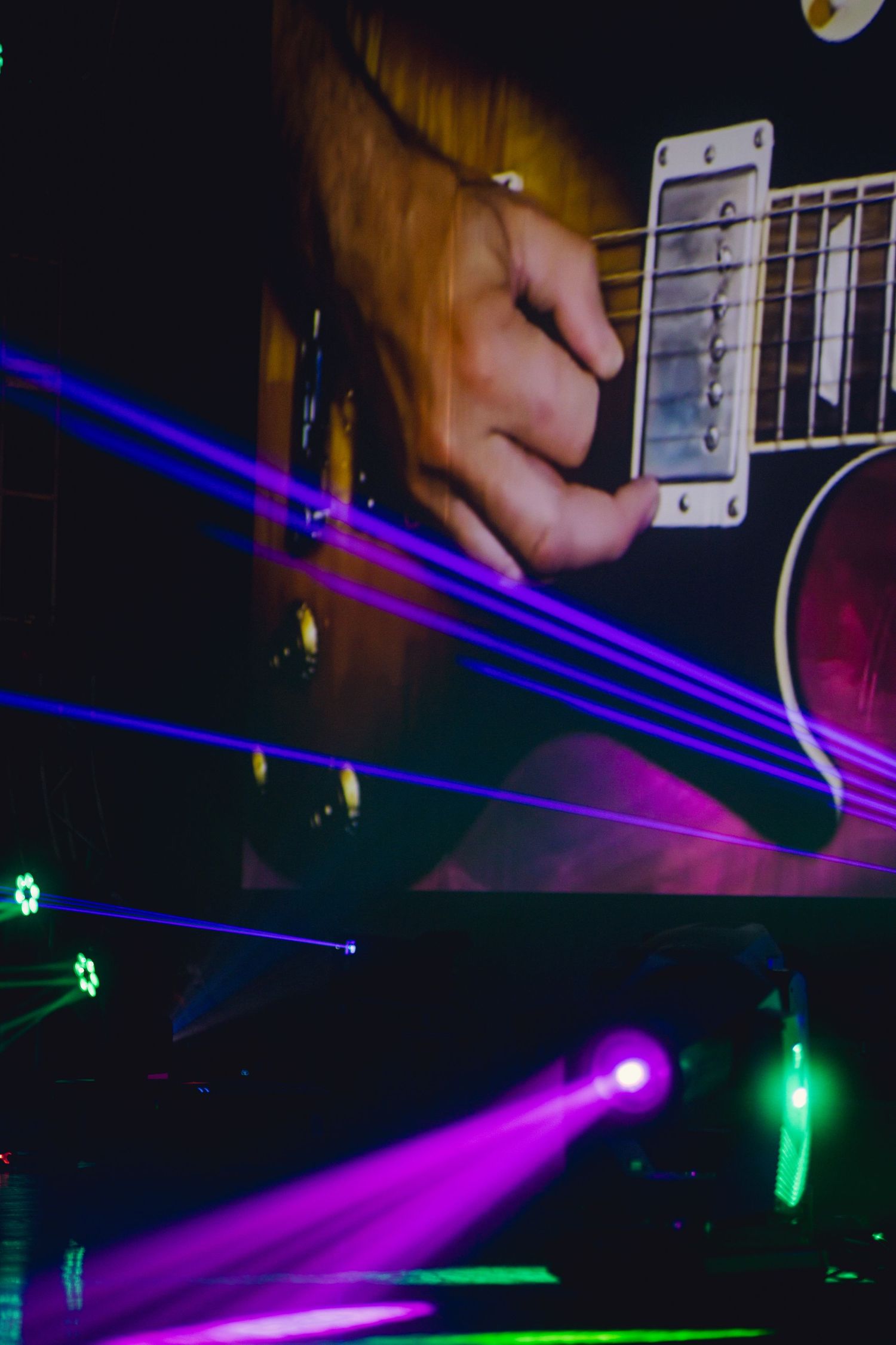 Electric guitar being played under colorful purple and green stage lighting with laser beam effects.