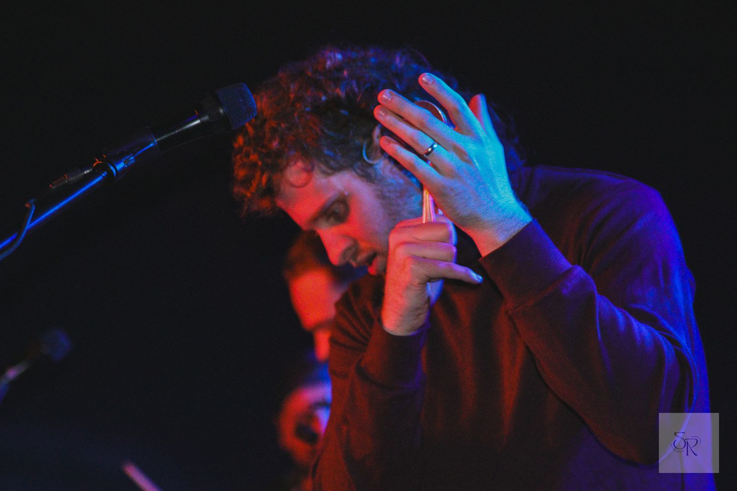 A performer sings passionately into a microphone under dramatic blue and red stage lighting during a live concert performance.