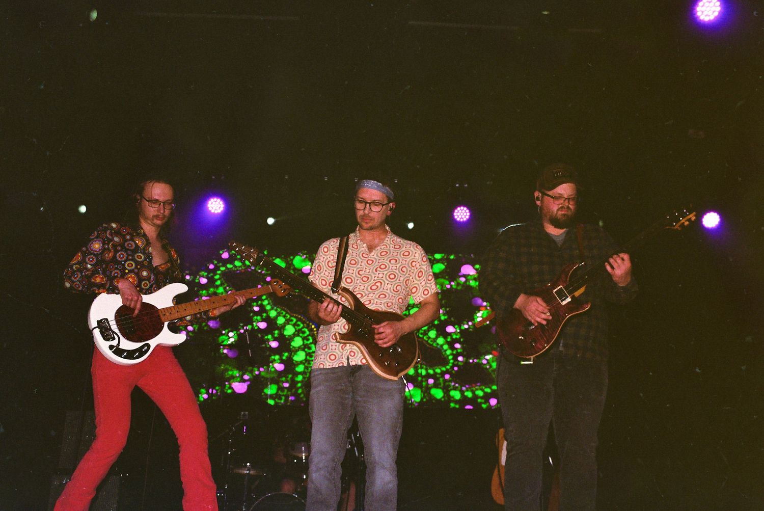 Three guitarists performing on stage with green holiday lights decorating the background.