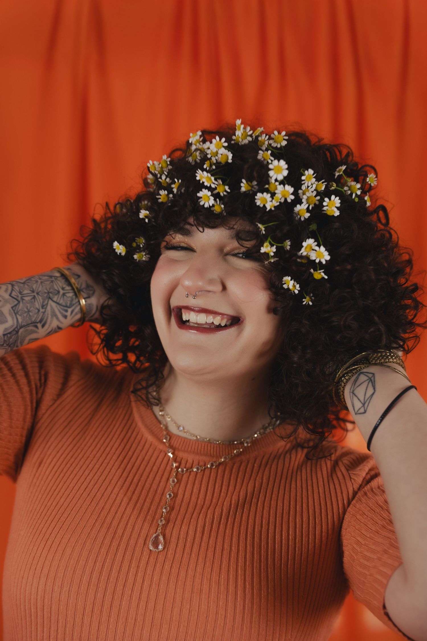 Smiling woman with curly hair adorned with daisy flowers, wearing an orange ribbed top and tattoos against an orange backdrop.