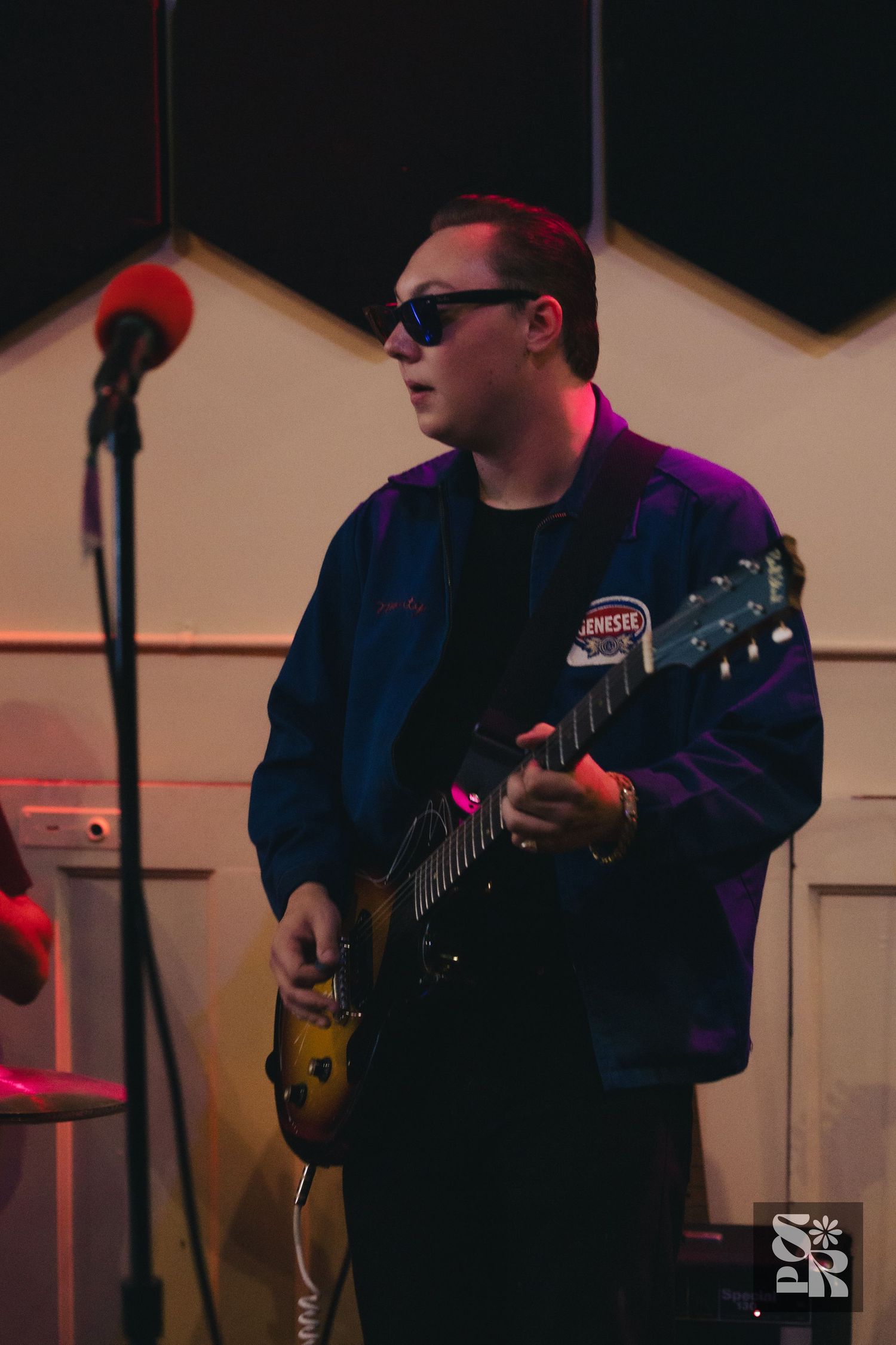 Musician in sunglasses and blue jacket playing electric guitar on stage with microphone stand nearby.