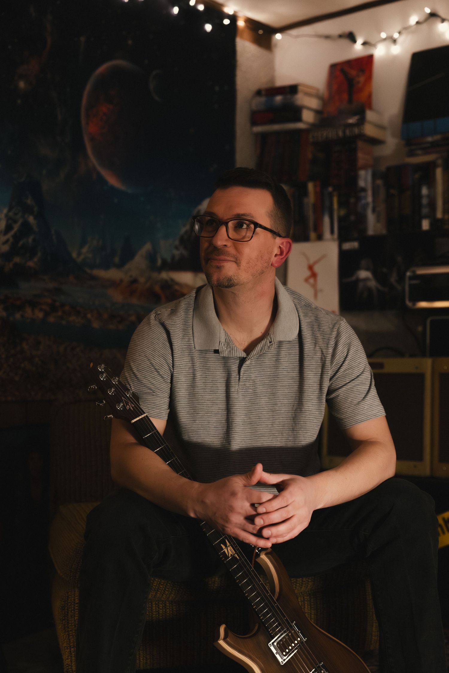 Man with glasses sitting with a gold electric guitar in a dimly lit music room with space-themed decor.