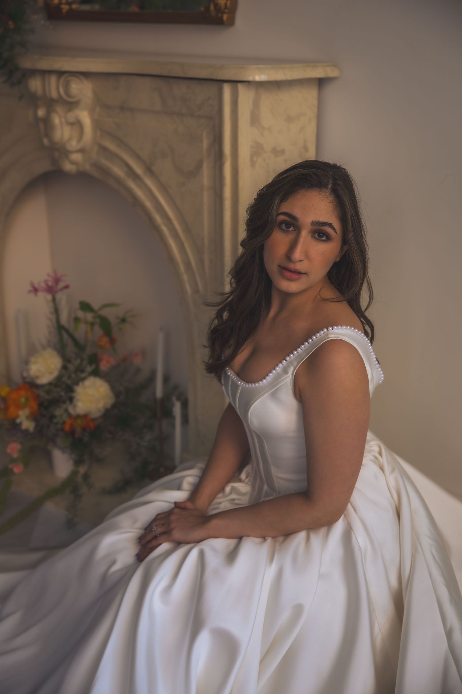 A bride in a classic white ballgown poses by an ornate fireplace with floral arrangements.