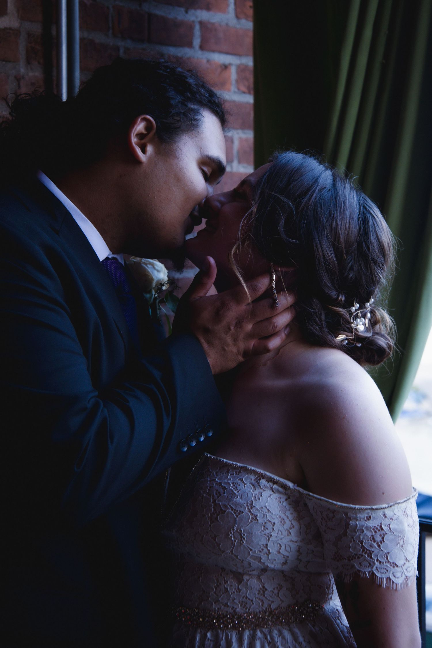 A romantic moment between newlyweds in formal attire against a brick wall and green curtain backdrop.